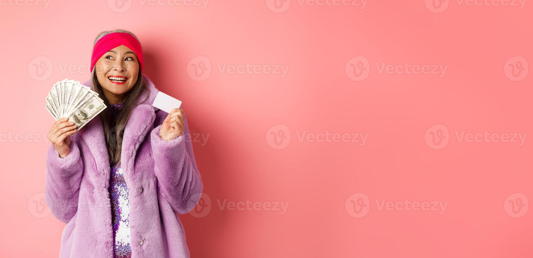 Shopping and fashion concept. Fashionable asian senior woman dreaming about stores and sales, holding dollars money and plastic credit card, pink background photo