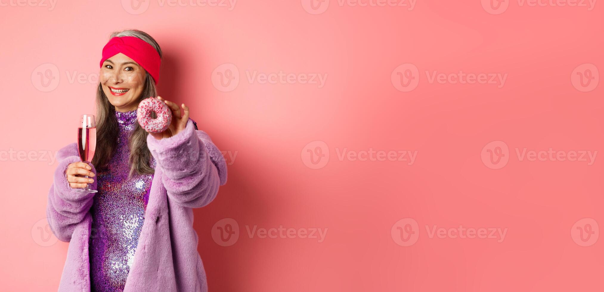 Cheerful asian elderly woman in fancy clothes drinking champagne, giving you donut, extend hand with dessert and smiling, pink background photo