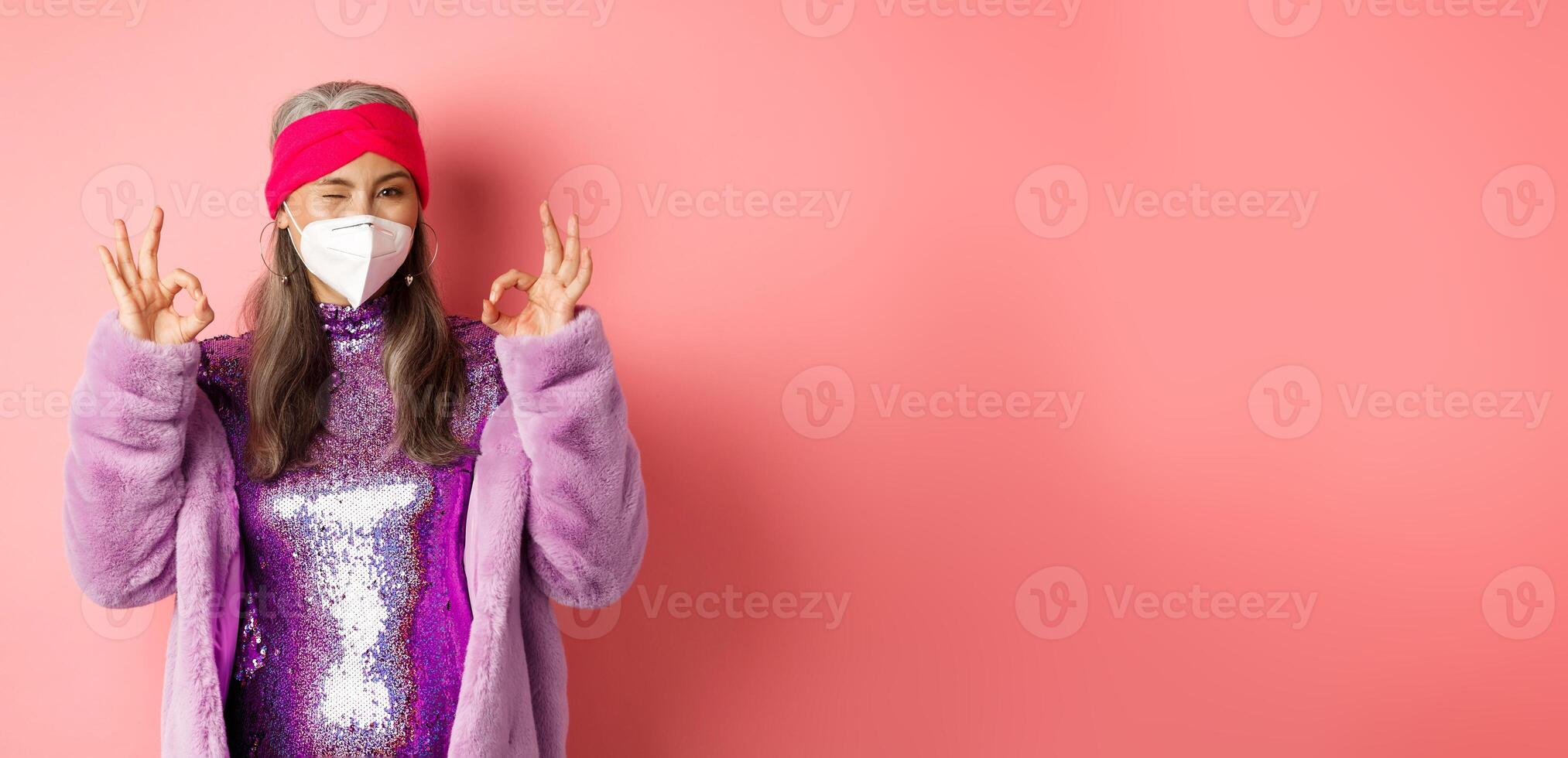 Covid-19, pandemic and fashion concept. Awesome asian grandmother wearing stylish disco dress and respirator, showing okay signs and winking, no proble gesture photo