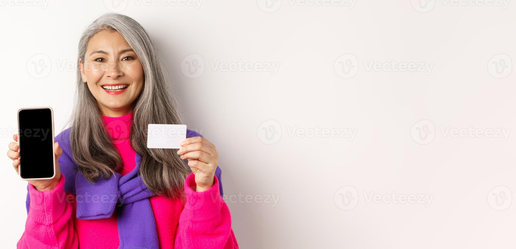 Online shopping. Closeup of smiling asian senior woman showing blank mobile screen and plastic credit card, looking happy, standing over white background photo