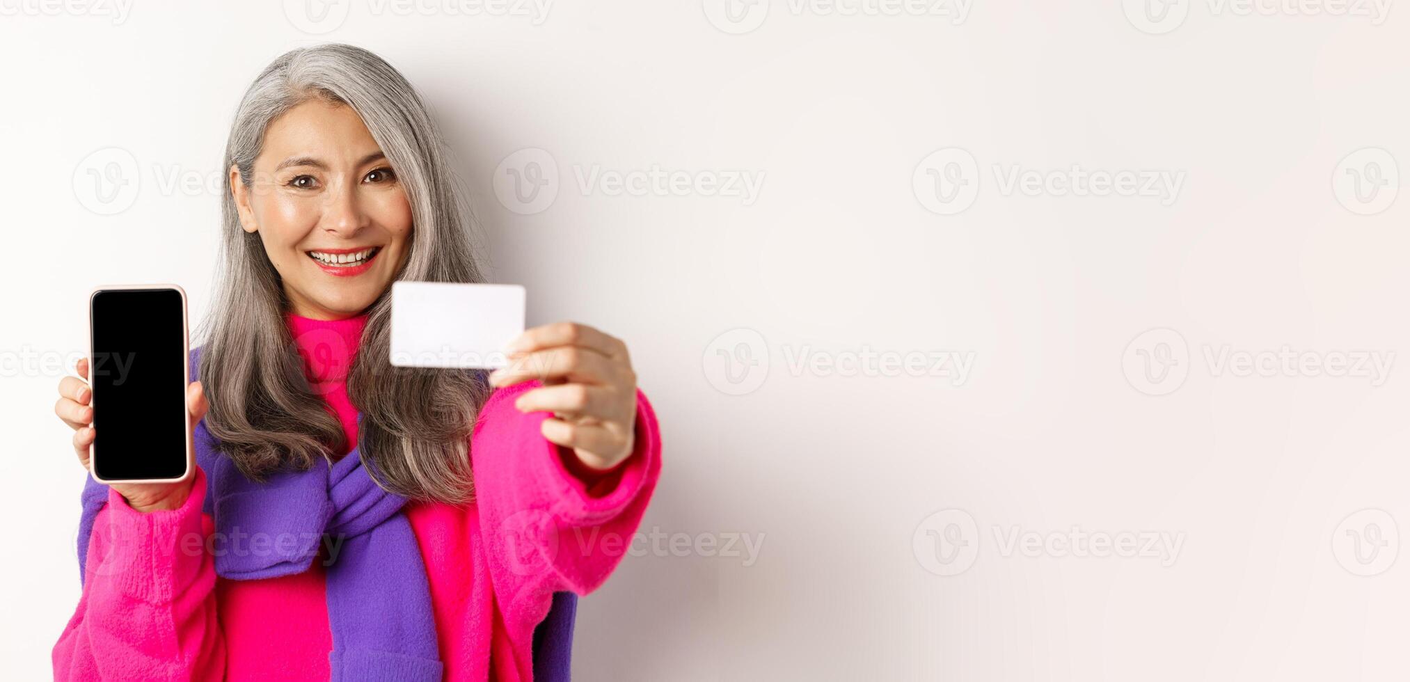 Online shopping. Close-up of stylish korean senior woman showing empty mobile screen and plastic credit card, smiling happy, standing over white background photo
