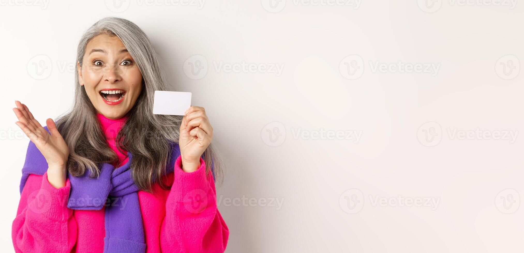 Shopping concept. Happy asian old aldy looking impressed and showing plastic credit card of her bank, standing over white background photo