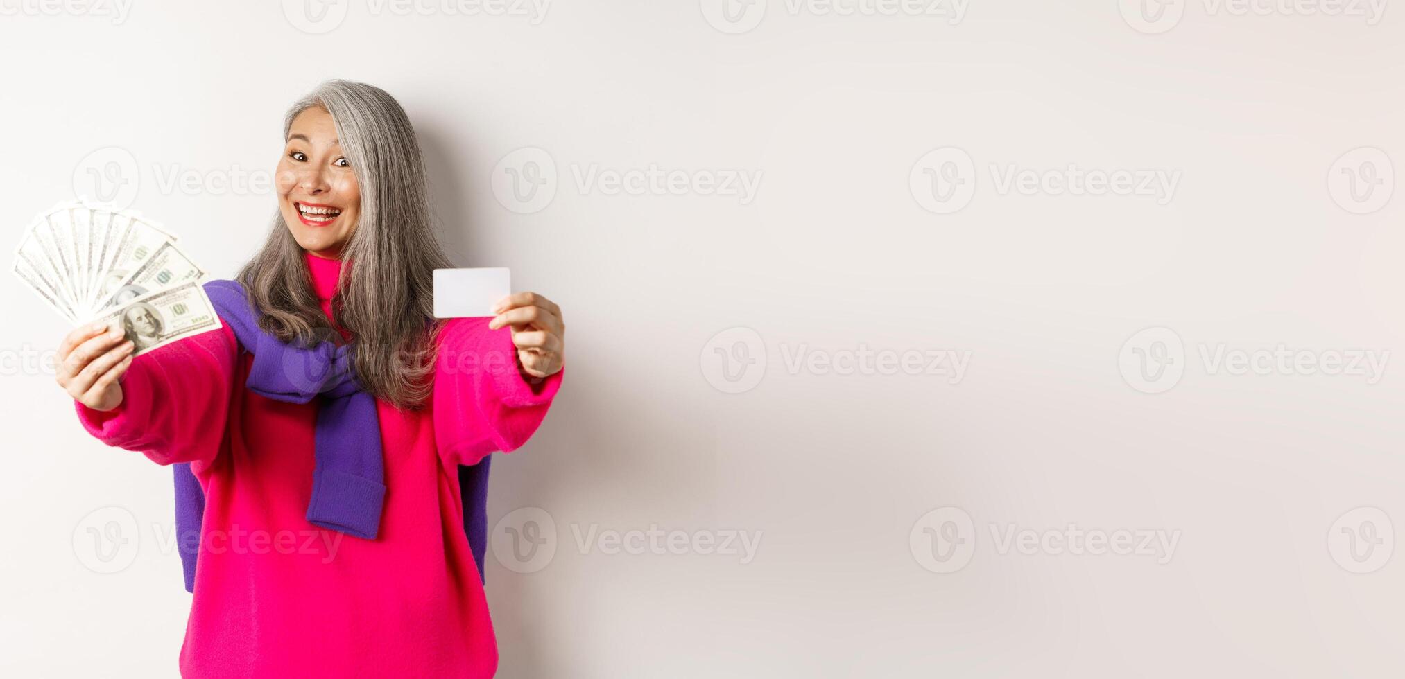 Shopping concept. Cheerful asian senior woman showing cash and plastic credit card, smiling at camera, standing over white background photo