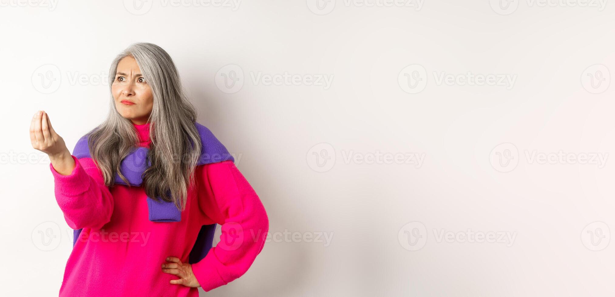 Grumpy asian grandmother complaining, showing italian what do you want gesture, shaking fingers and staring angry left at copy space, standing over white background photo
