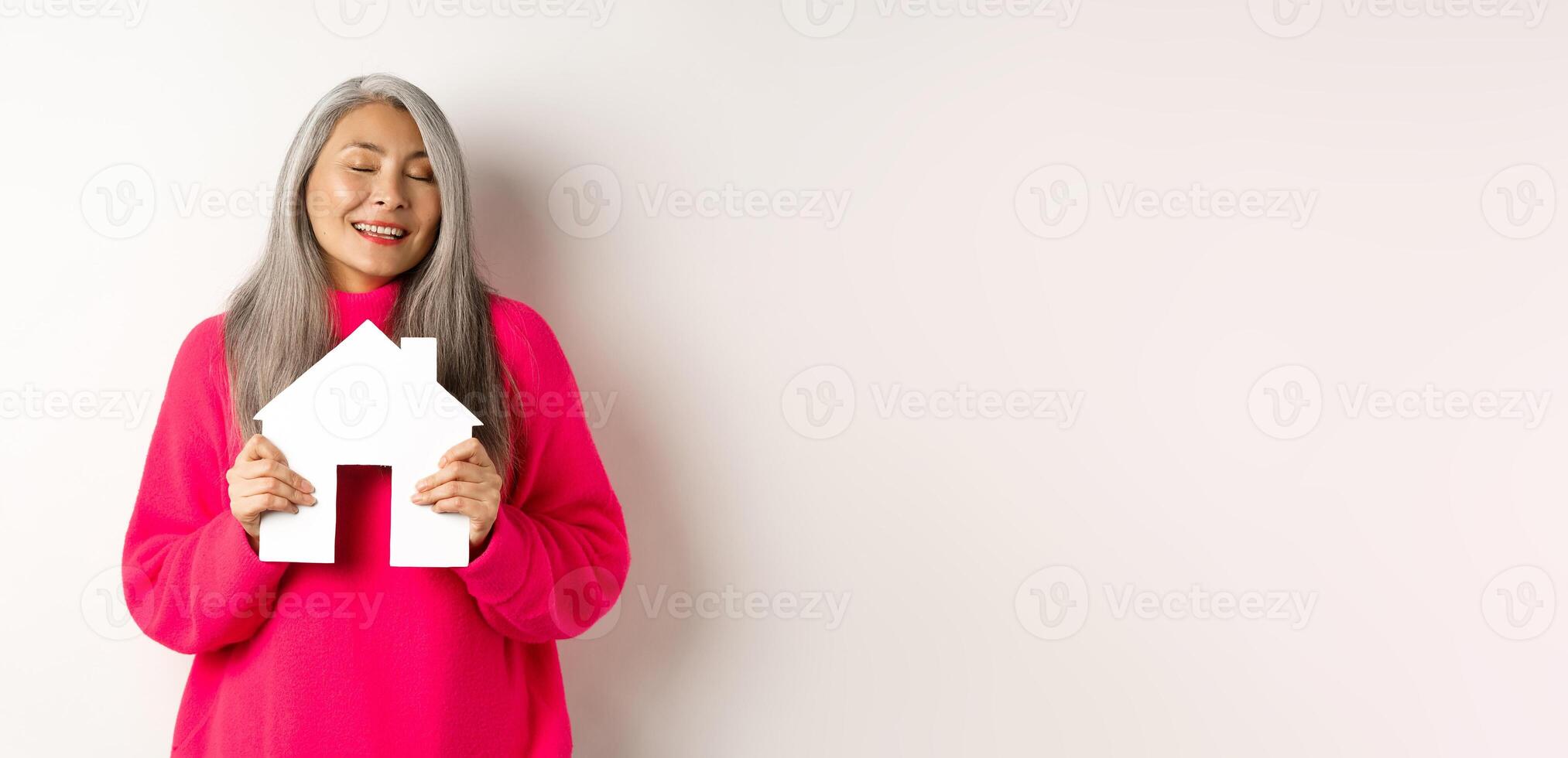 Real estate. Beautiful dreamy asian lady hugging paper house model with closed eyes, smiling as dreaming about buying apartment, standing over white background photo