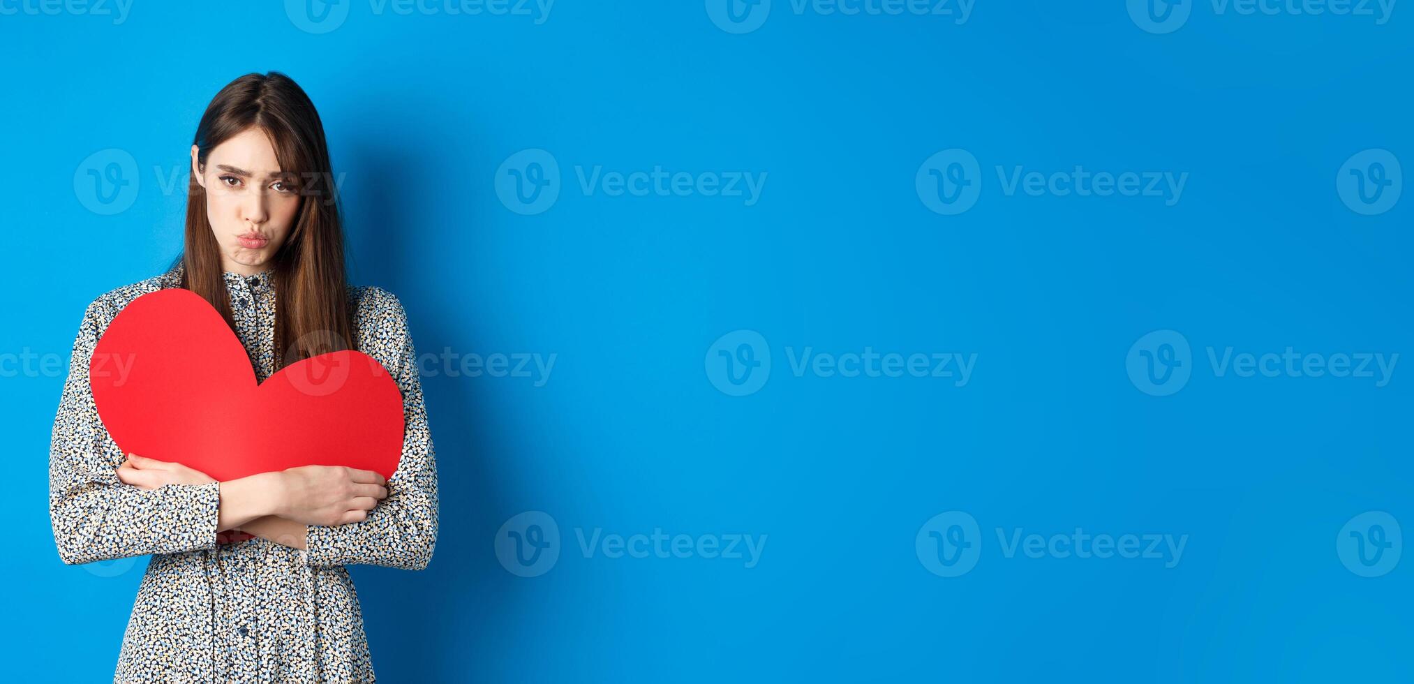 Valentines day. Sad and gloomy caucasian girl pucker lips and looking disappointed, holding big red heart cutout, standing on blue background photo