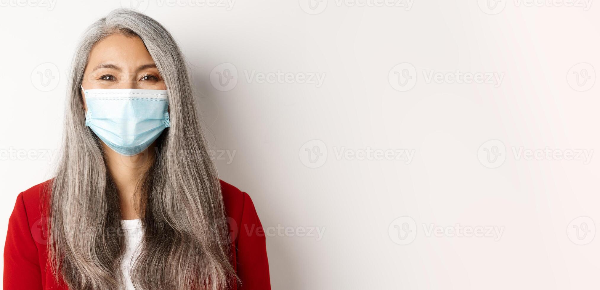 Covid, pandemic and business concept. Close-up of cheerful asian senior businesswoman in medical mask smiling at camera, white background photo
