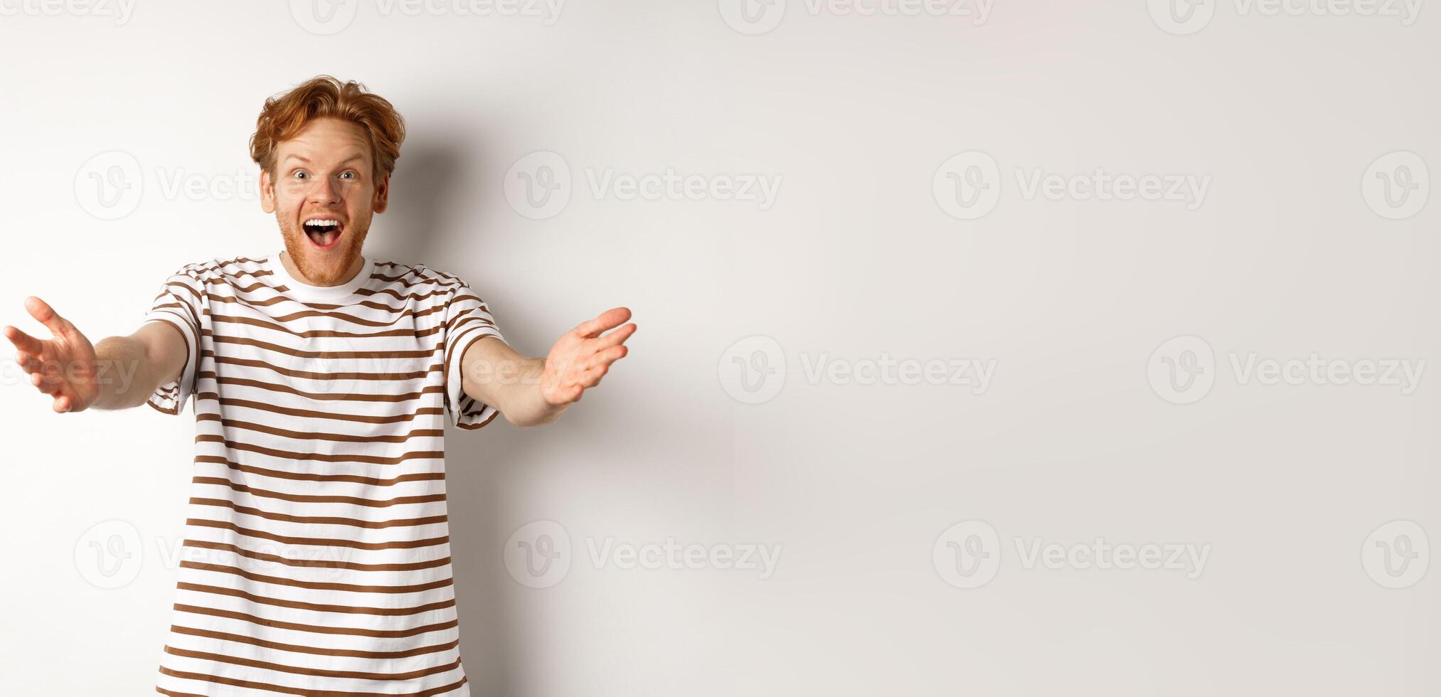 Cheerful man with red curly hair reaching hands forward, stretch out arms to welcome or congratulate you, smiling happy, standing over white background photo