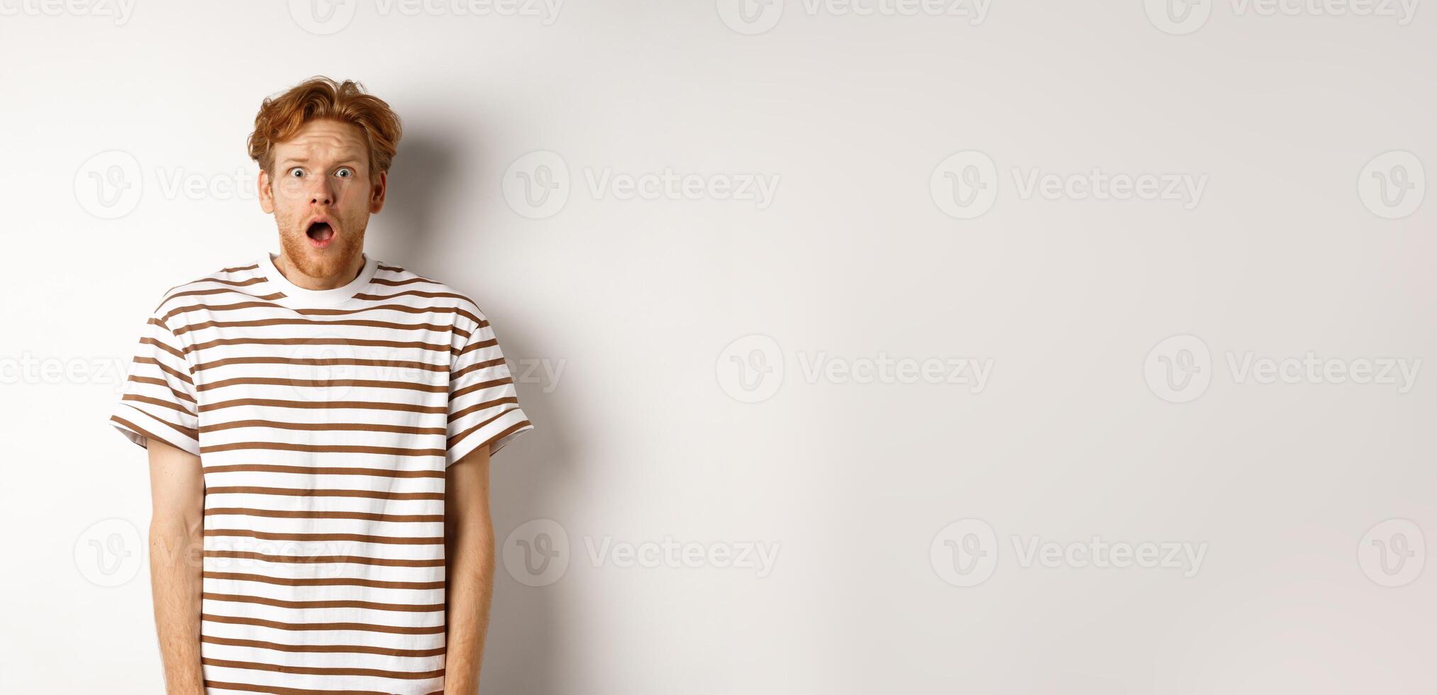 Shocked and scared redhead man screaming, open mouth and staring terrified at camera, standing over white background photo