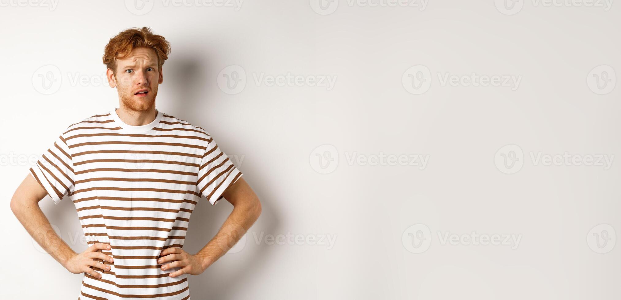Confused and offended man with ginger curly hair staring at camera puzzled, standing questioned against white background photo
