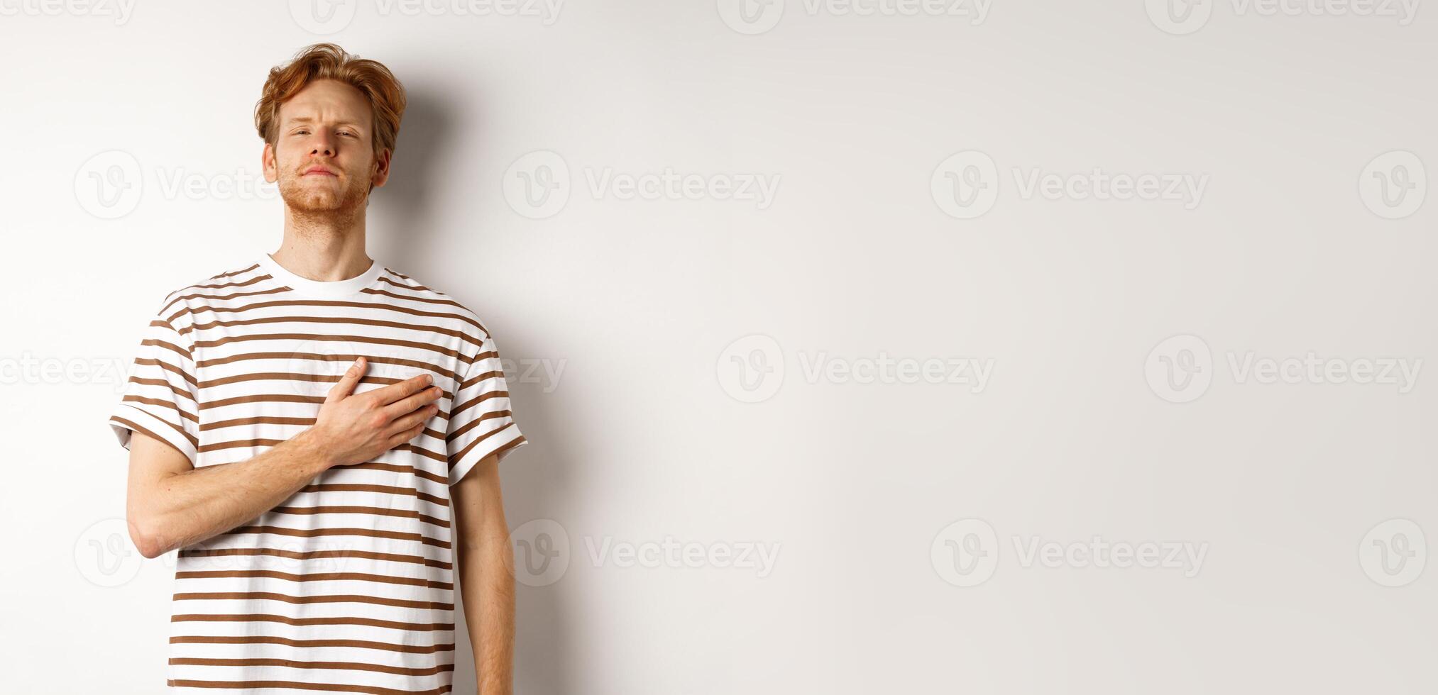 Young bearded redhead man holding hand on heart and looking patriotic, standing during national anthem, standing over white background photo