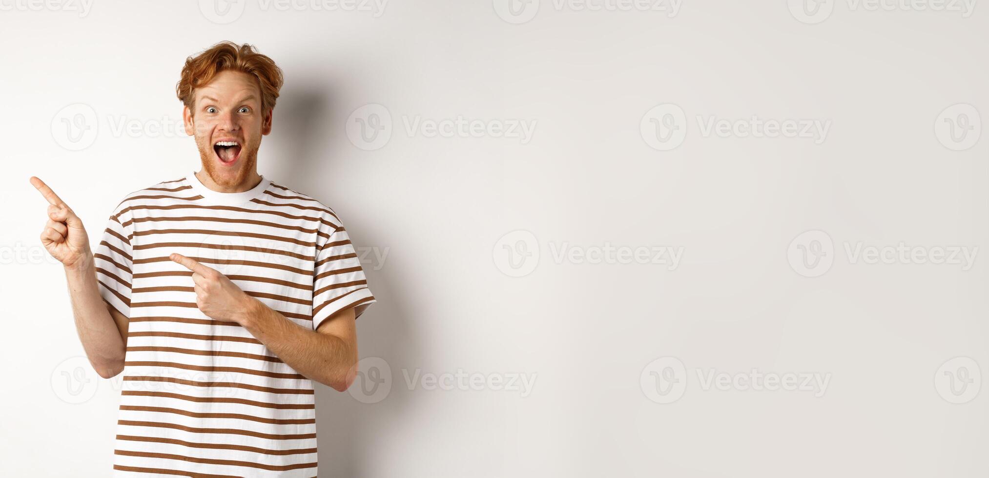 Cheerful man with red hair checking out promo offfer, pointing left at logo and smiling at camera, standing over white background photo