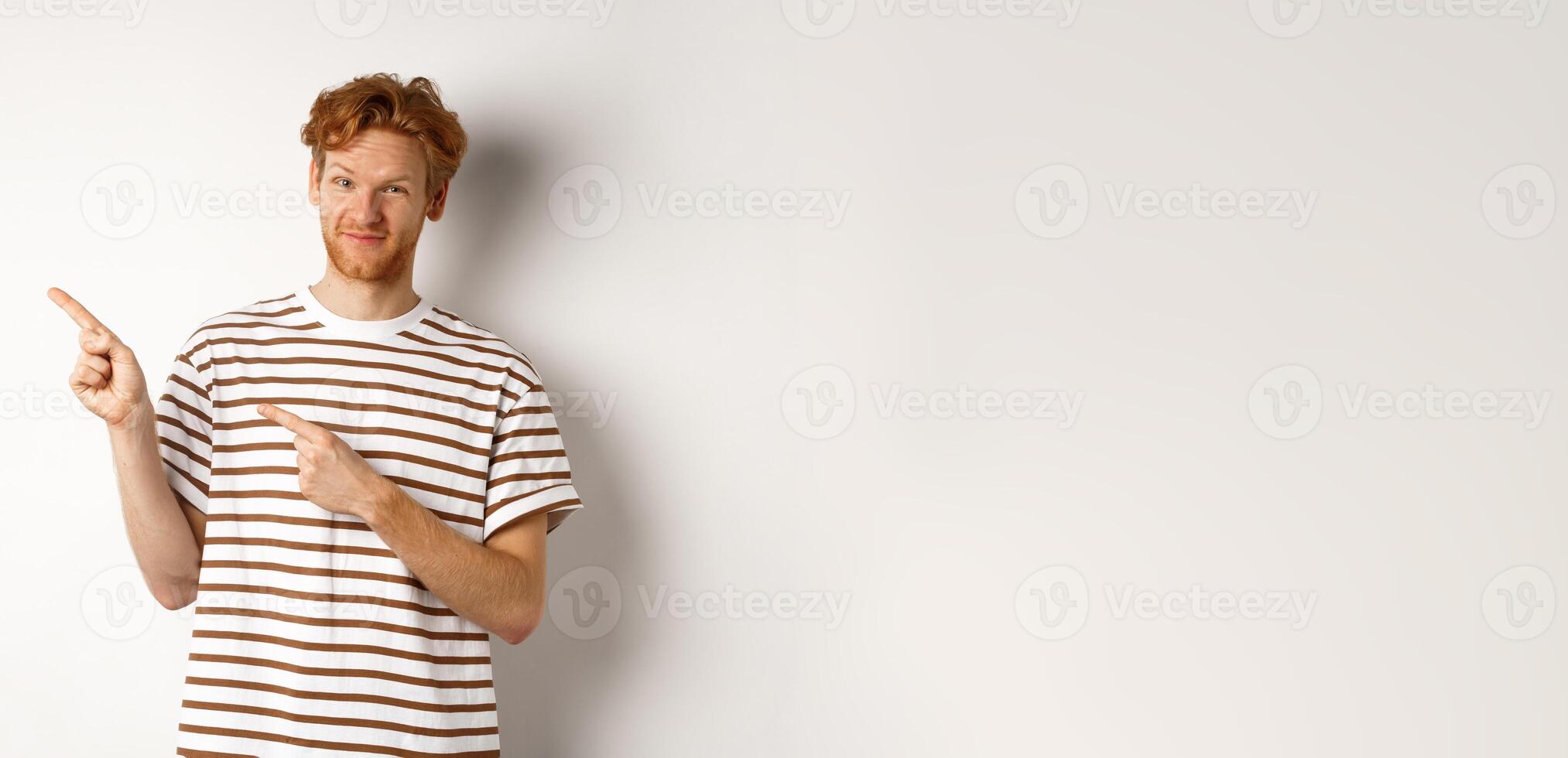 Doubtful redhead guy pointing fingers left at looking skeptical at camera, standing over white background photo