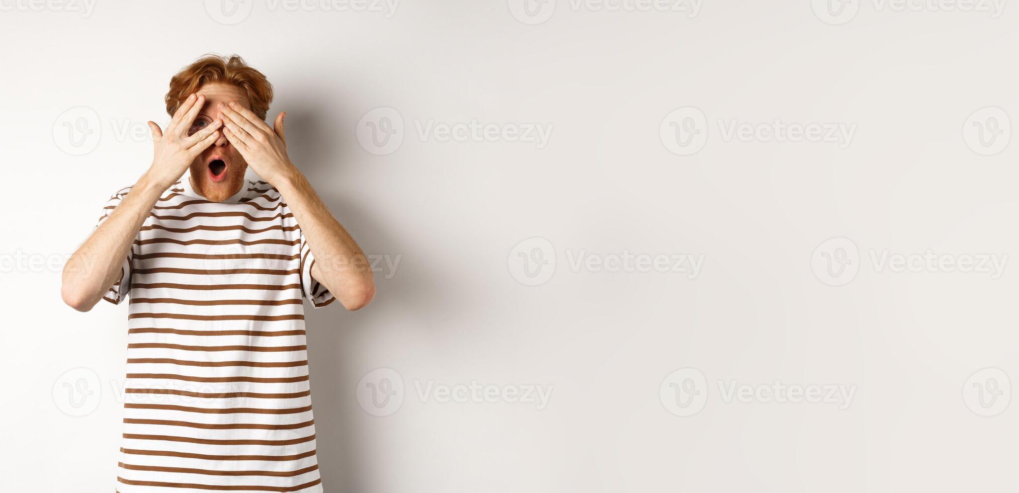 Shocked and embarrassed redhead guy covering eyes, peeking through fingers and gasping, standing over white background photo