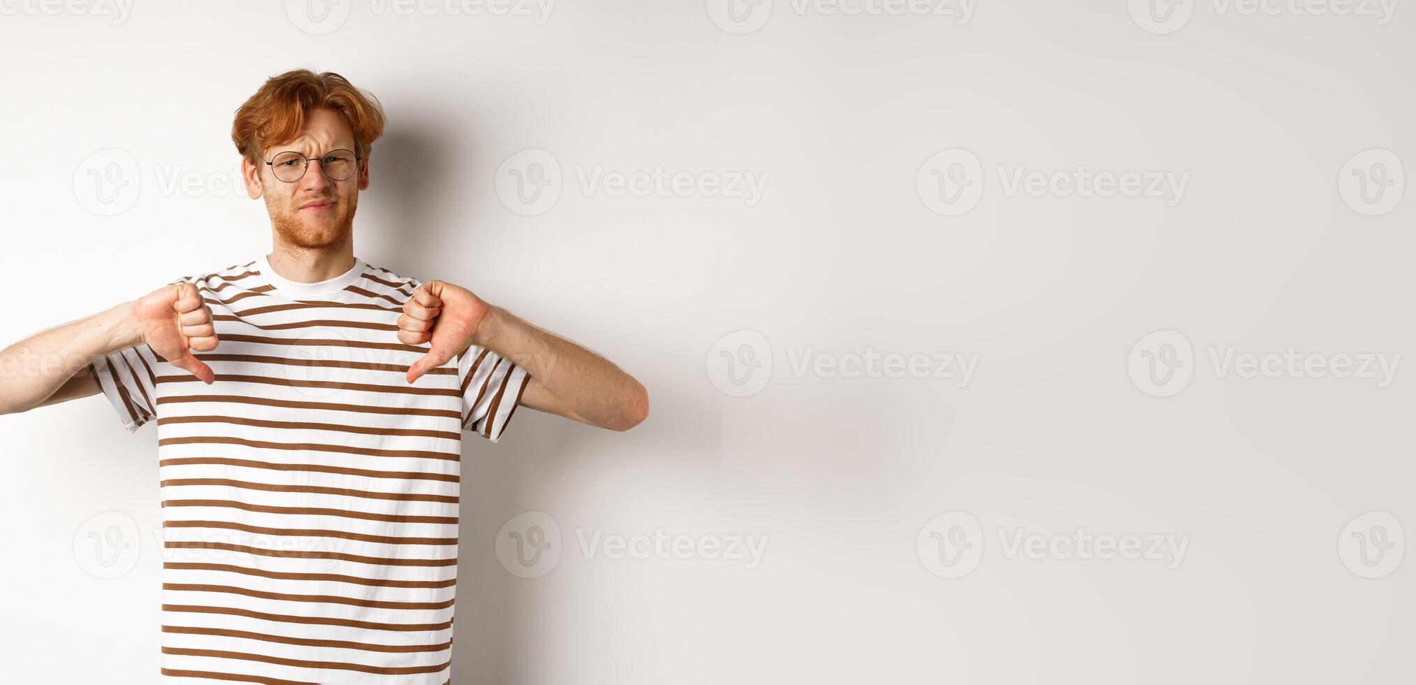 Disappointed young man with red hair and glasses disagree, showing thumbs-down and frowning displeased, standing over white background photo