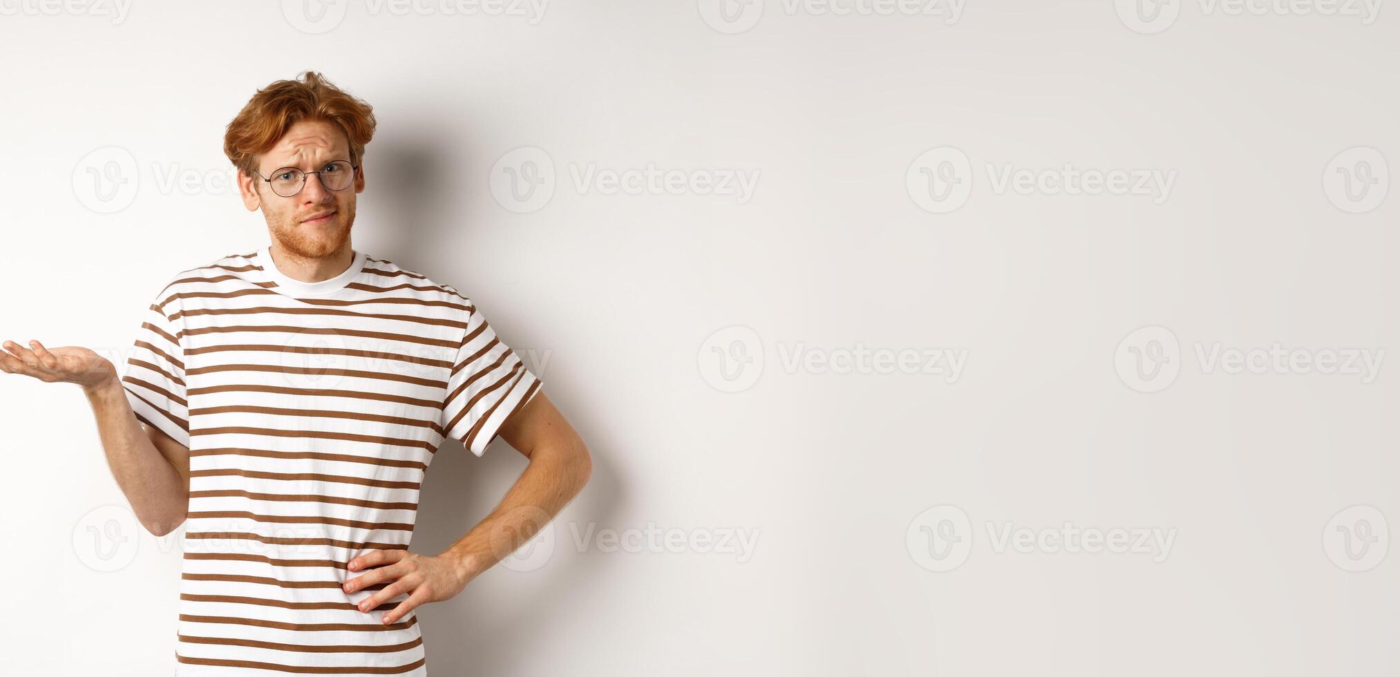 So what. Confused and annoyed redhead man waiting for reply, standing questioned with raised hand, holding something on palm, white background photo