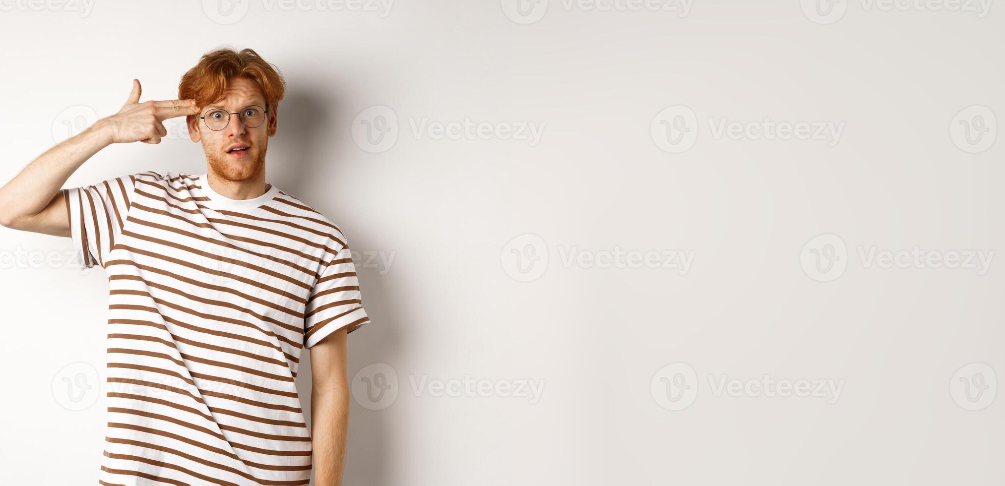 Annoyed teenage redhead guy making finger gun near head, staring bothered and irritated at camera, white background photo