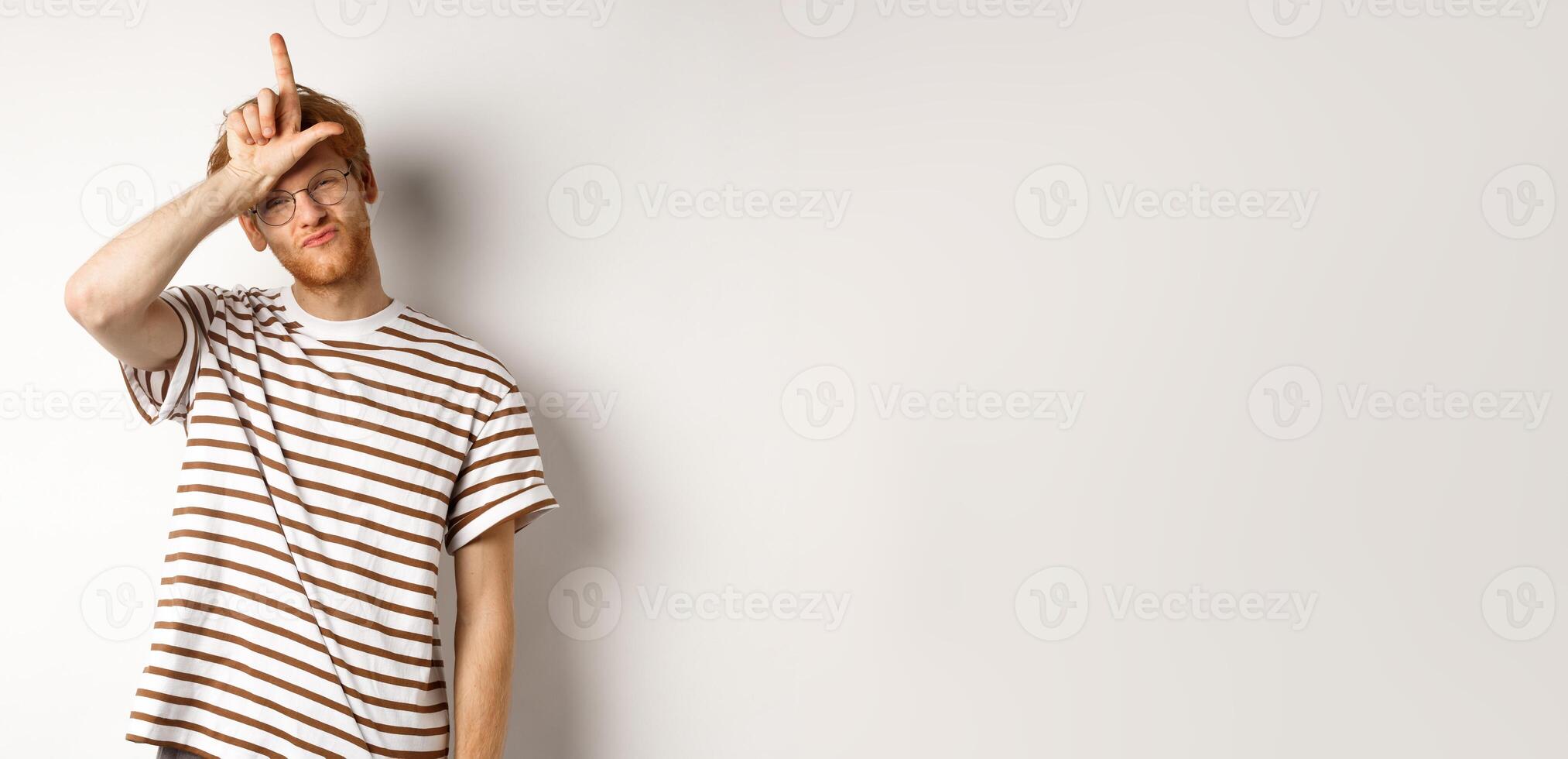 Sassy and arrogant redhead man scolding lost team, showing loser gesture on forehead and making smug face, standing over white background photo