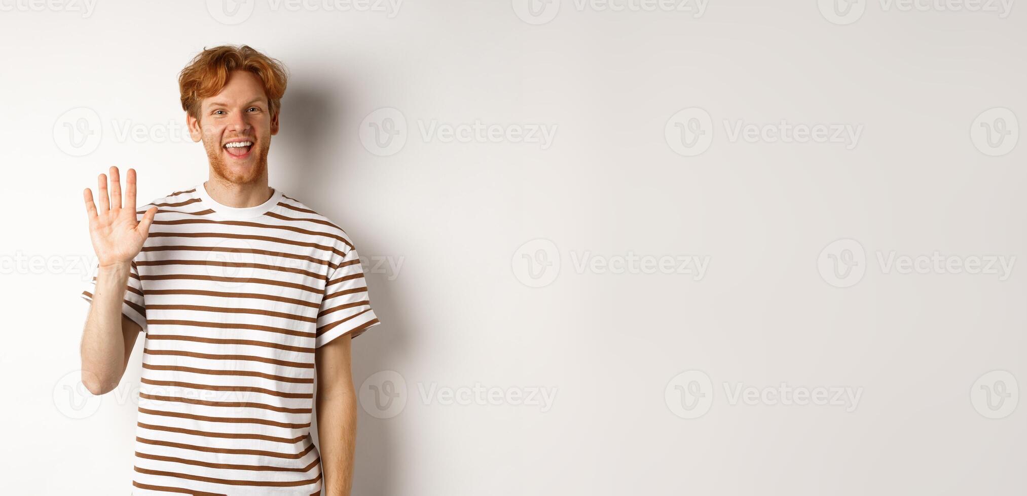 Friendly man with red hair and beard saying hi, waving hand and smiling, standing over white background photo