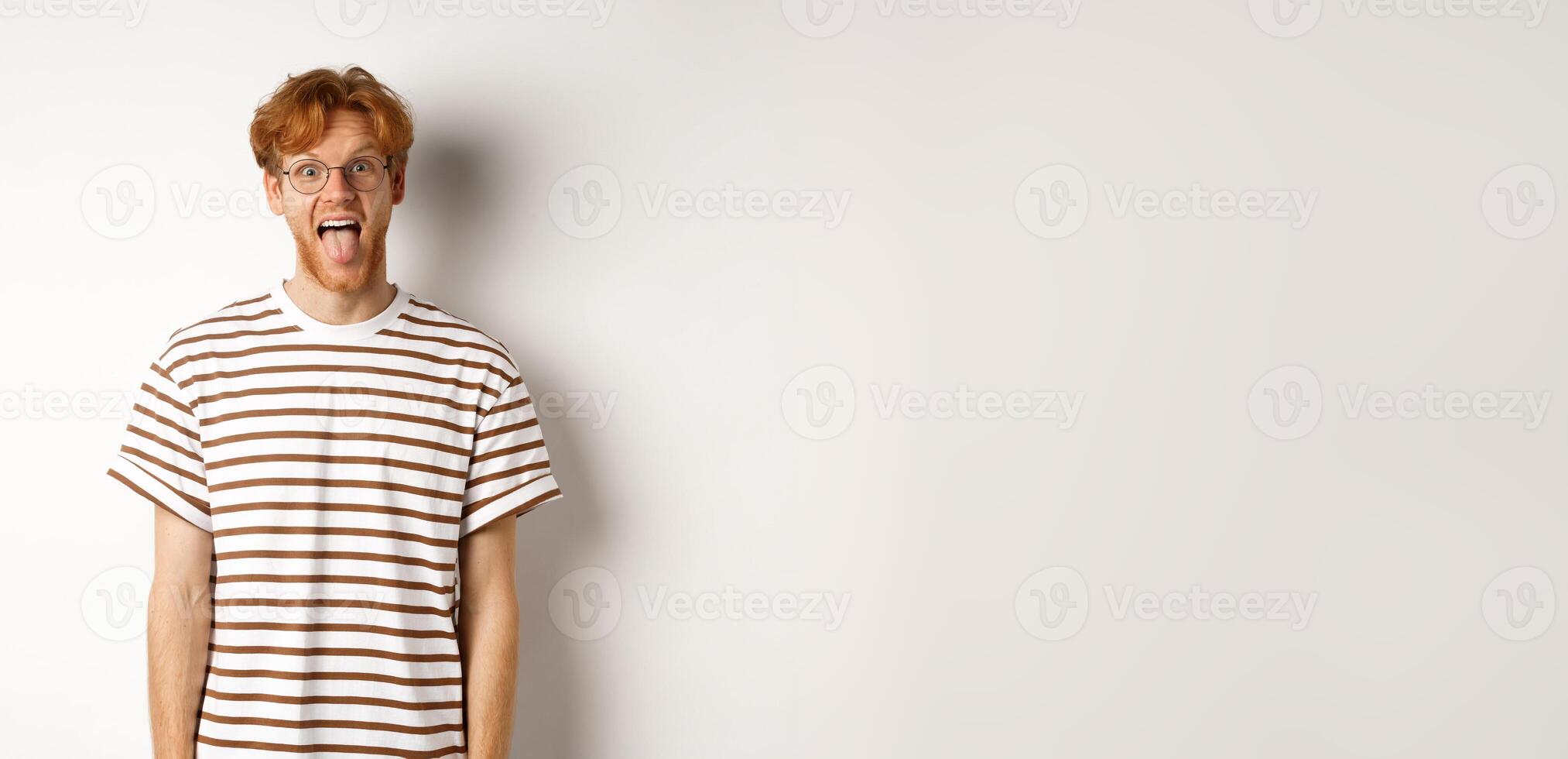 Funny young man with messy red hair and glasses showing tongue, staring at camera, standing over white background photo