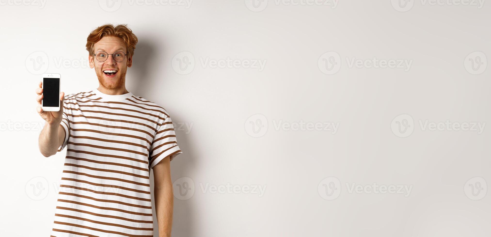 Technology and e-commerce concept. Happy young redhead man in glasses showing blank smartphone screen, looking at camera amazed, standing over white background photo
