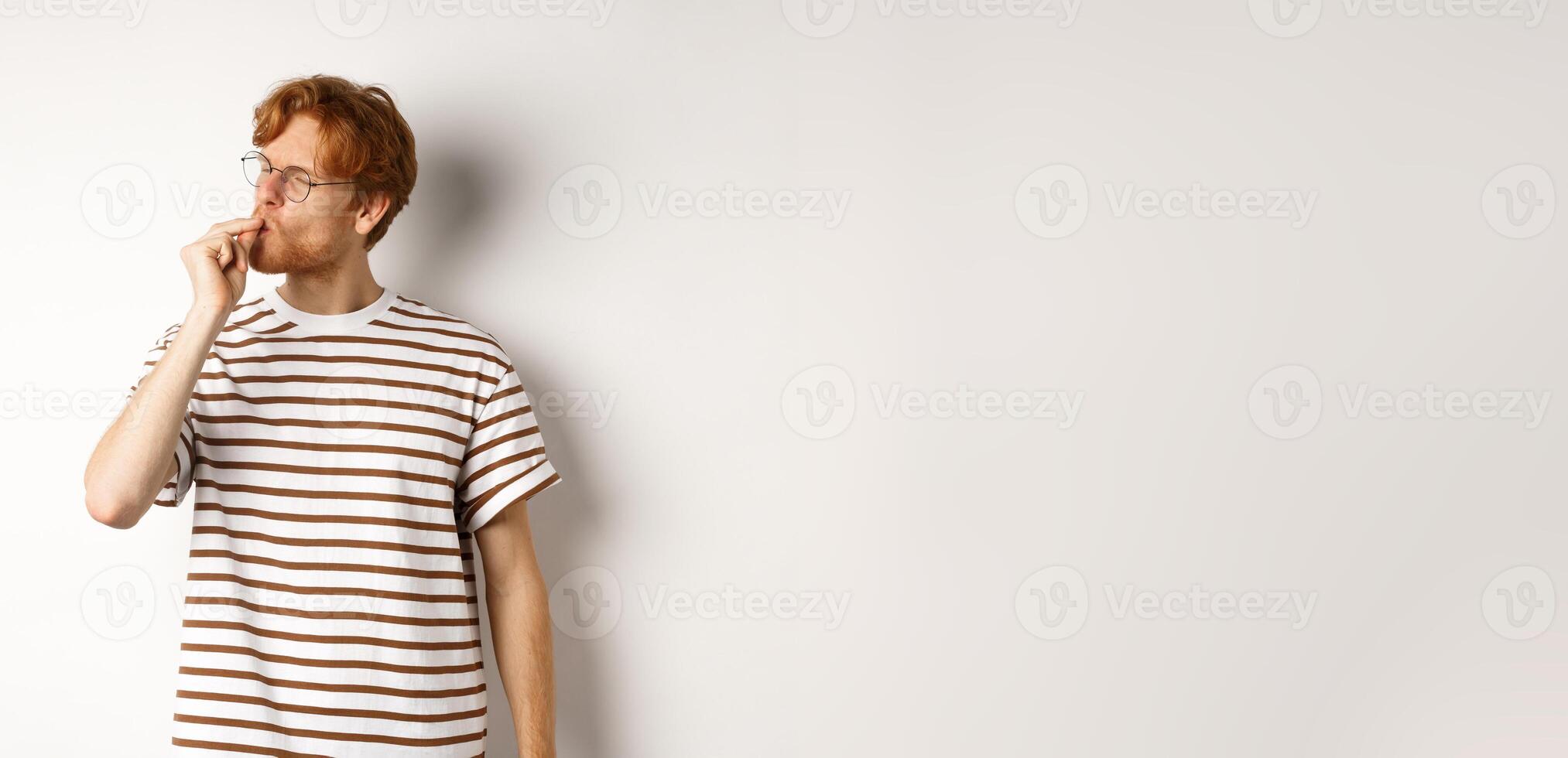 Image of satisfied young man with red hair and glasses kissing fingers, showing chefs kiss gesture to praise something perfect, standing over white background photo