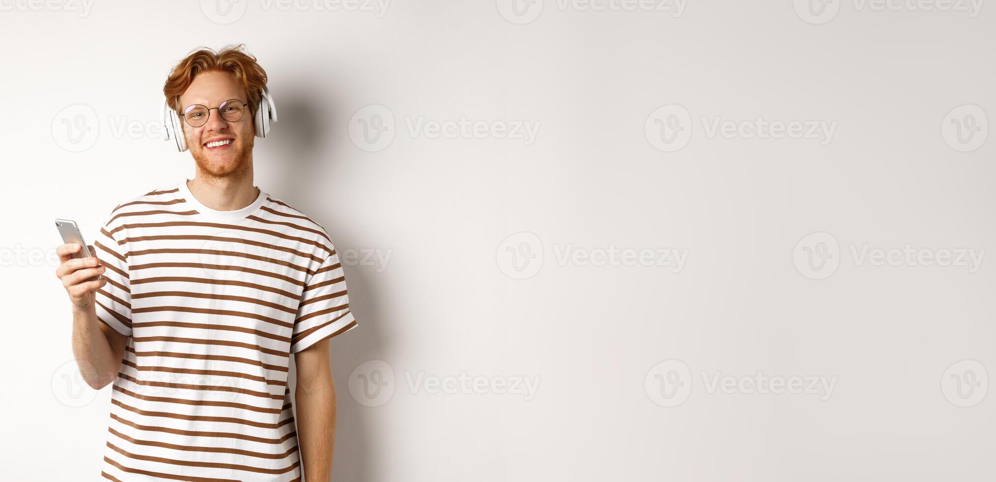 Technology concept. Young man with red hair and beard listening music in headphones and using smartphone, smiling at camera, white background photo