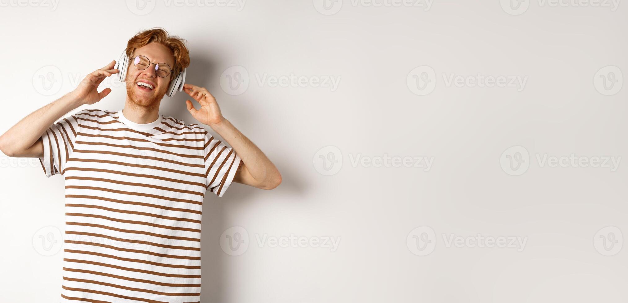 Technology concept. Happy redhead man listening music in headphones and singing along, standing over white background photo