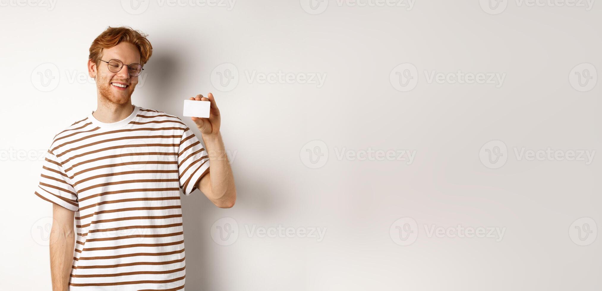 Shopping and finance concept. Cheerful redhead male student in glasses showing plastic credit card and looking satisfied, standing over white background photo