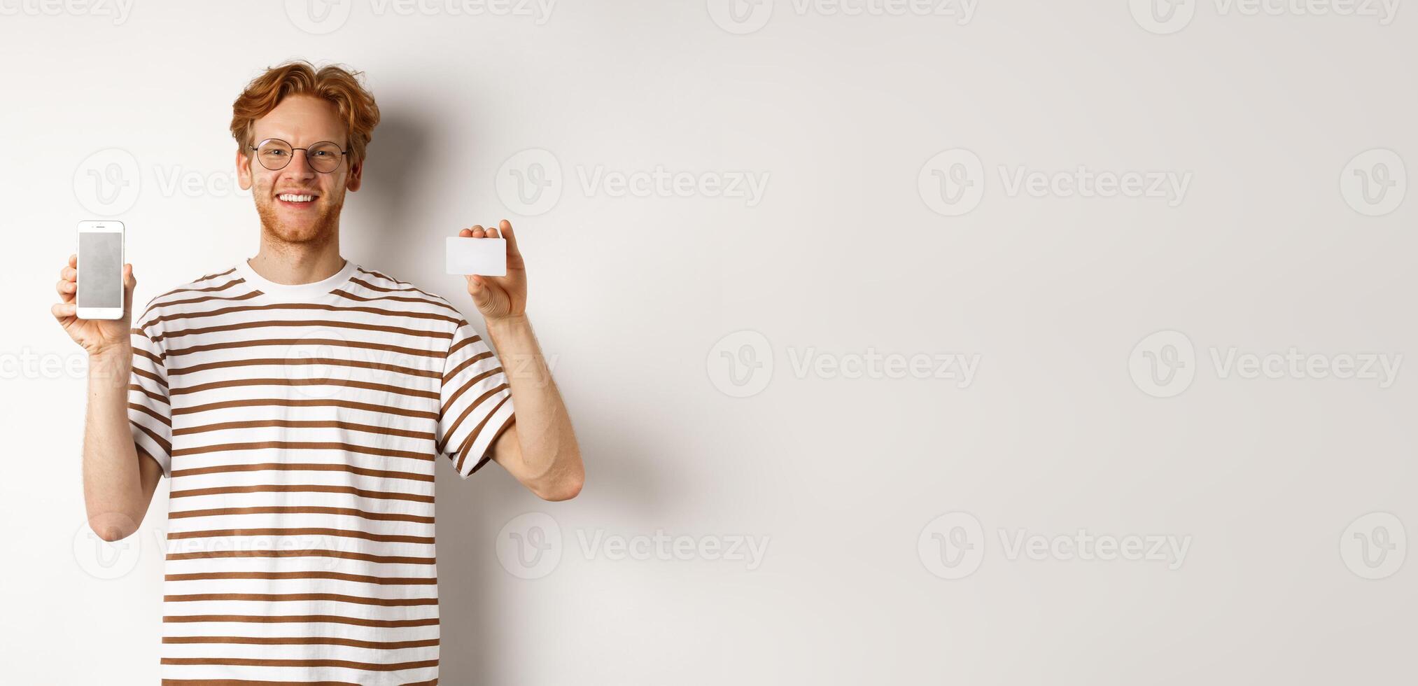 Shopping and finance concept. Young man showing blank mobile screen and plastic credit card, smiling at camera, white background photo
