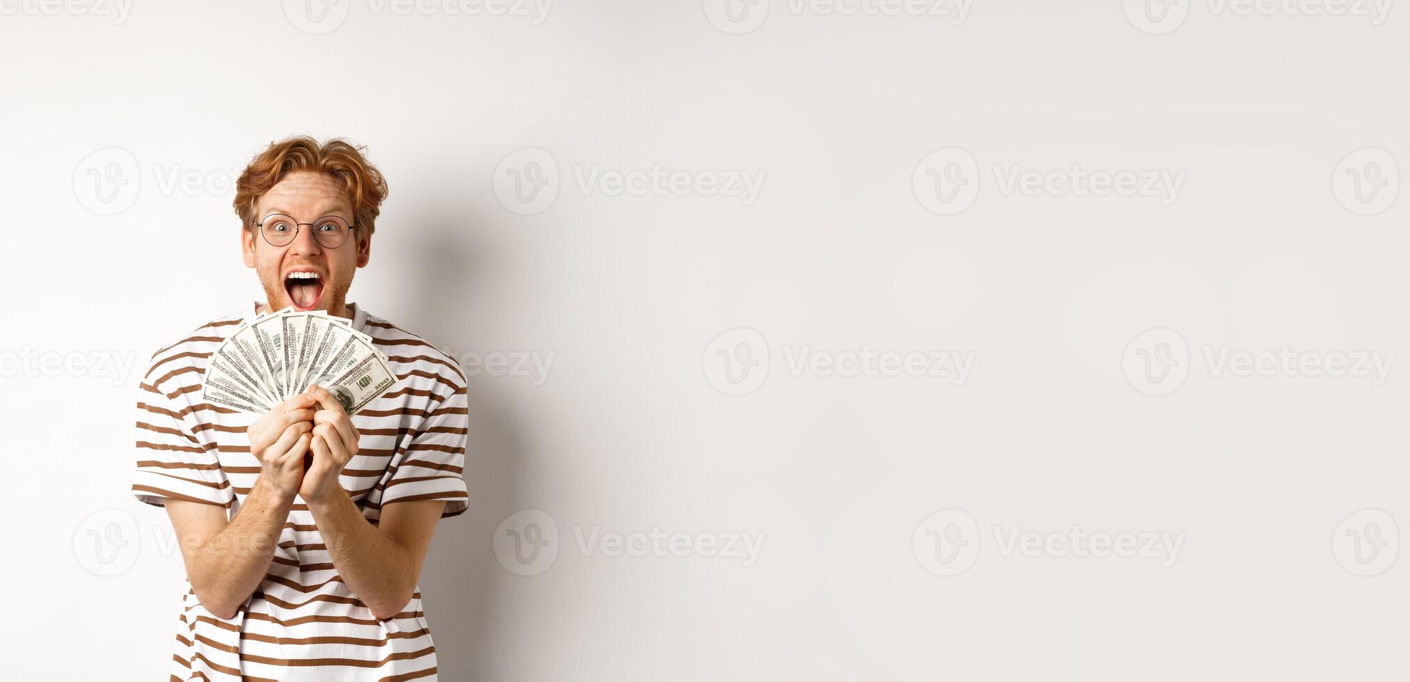 Lucky young man with red hair showing dollars, winning money and screaming of happiness, holding prize cash, standing over white background photo