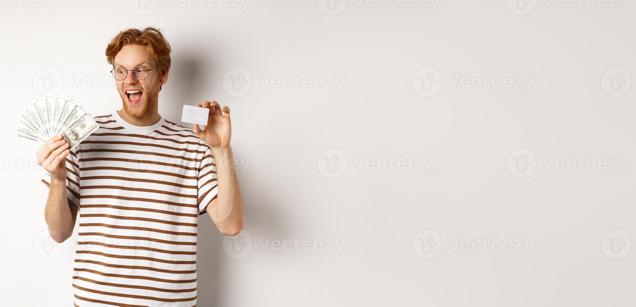 Shopping and finance concept. Cheerful handsome redhead man showing plastic credit card and cash, smiling amazed, white background photo