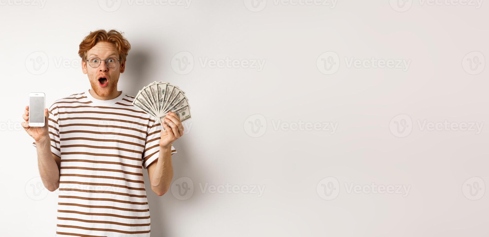 Amazed redhead man showing smartphone app on blank screen and money, winning prize cash online, standing over white background photo