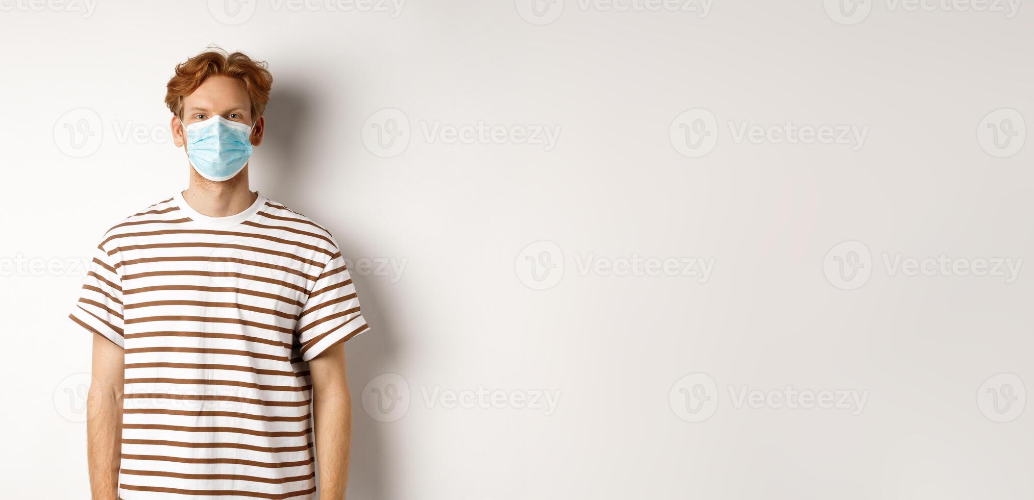 Covid-19, pandemic and social distancing concept. Young man with red hair wearing medical mask to prevent catching coronavirus, white background photo