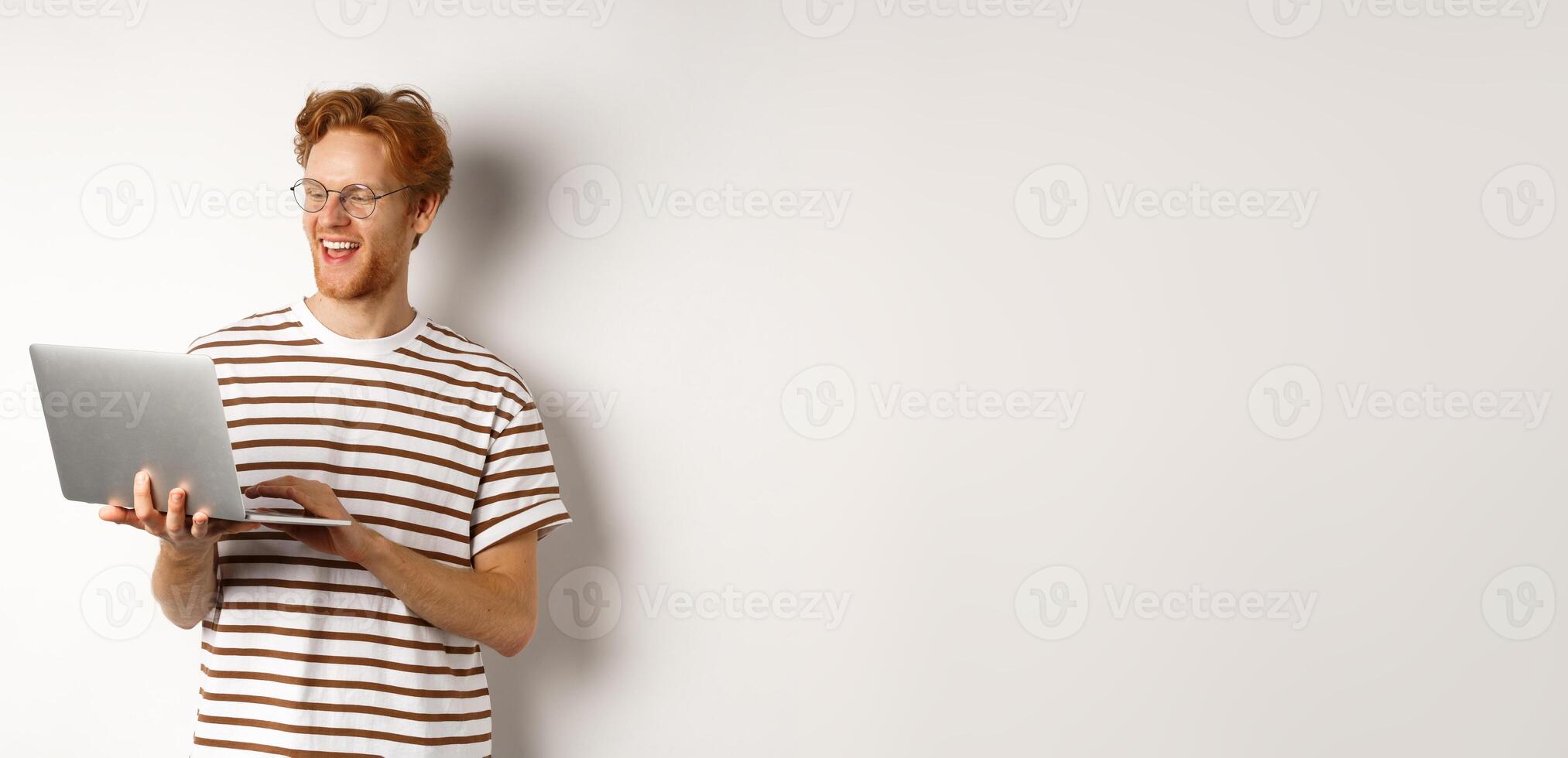 Handsome redhead man working freelance on laptop, smiling and looking pleased at computer screen, white background photo