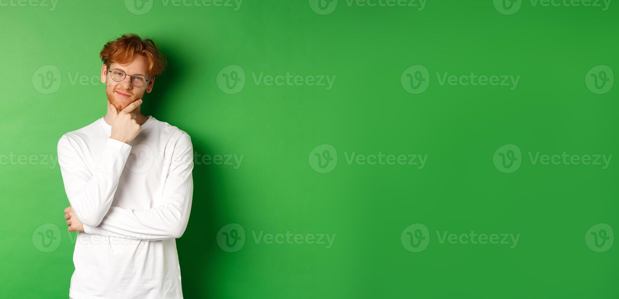 Intrigued young man with red hair, wearing glasses, looking at camera pleased and thoughtful, having an idea, standing over green background photo