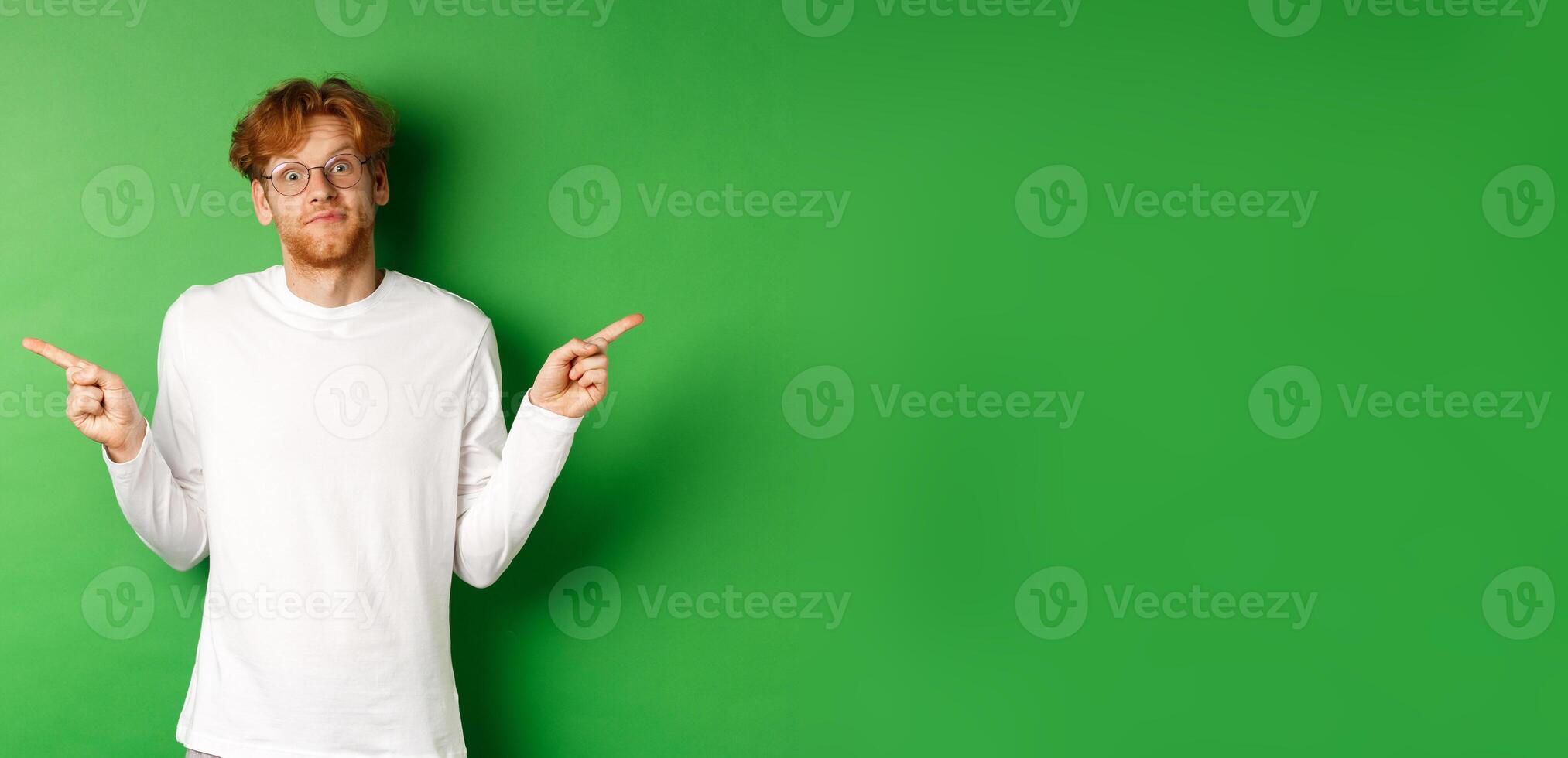 Indecisive funny guy with red hair pointing fingers sideways, staring confused at camera and showing two promo offers, standing over green background photo