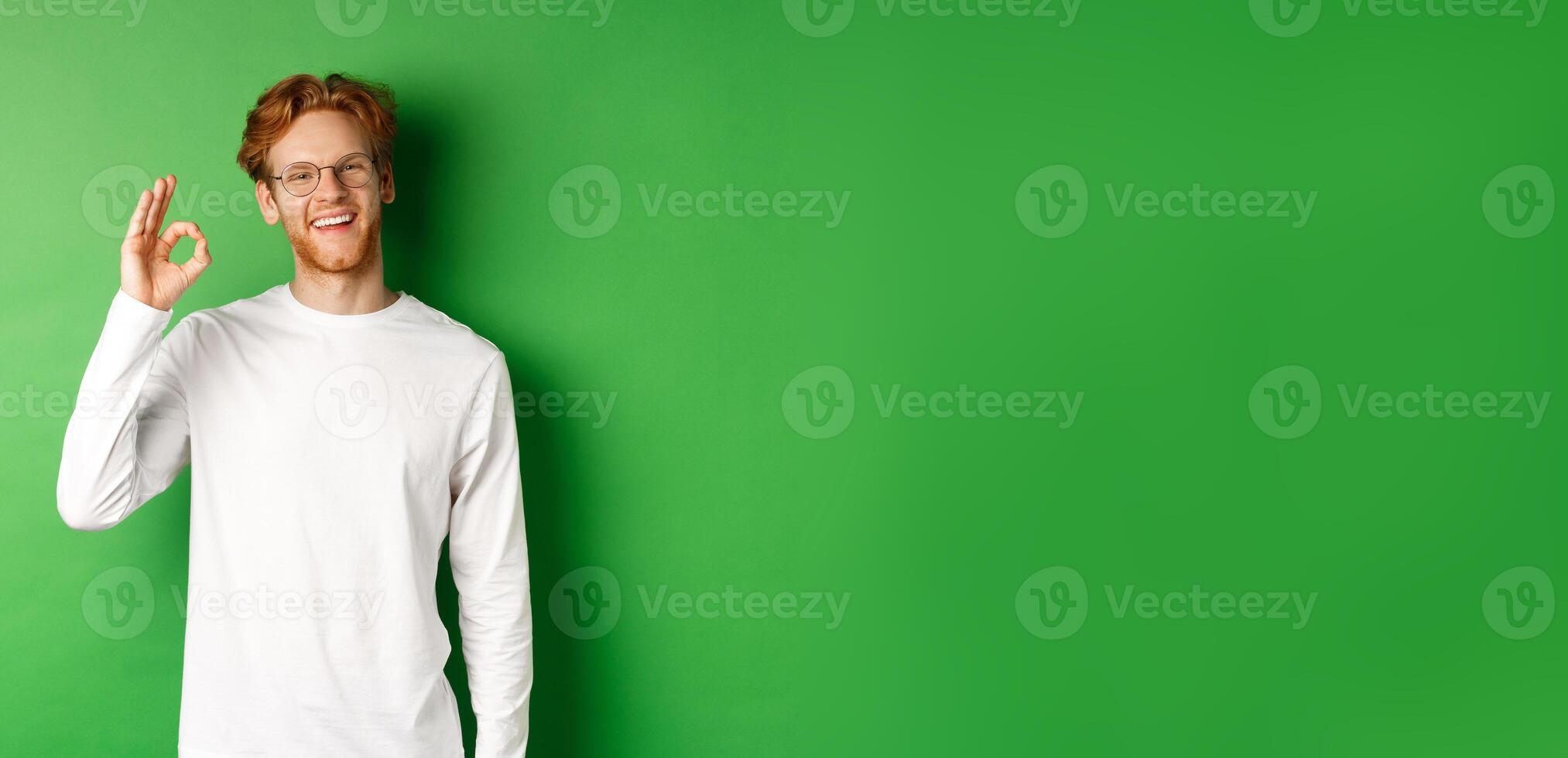 Happy male student with red hair, wearing glasses, showing okay sign in approval and smiling satisfied, standing over green background photo