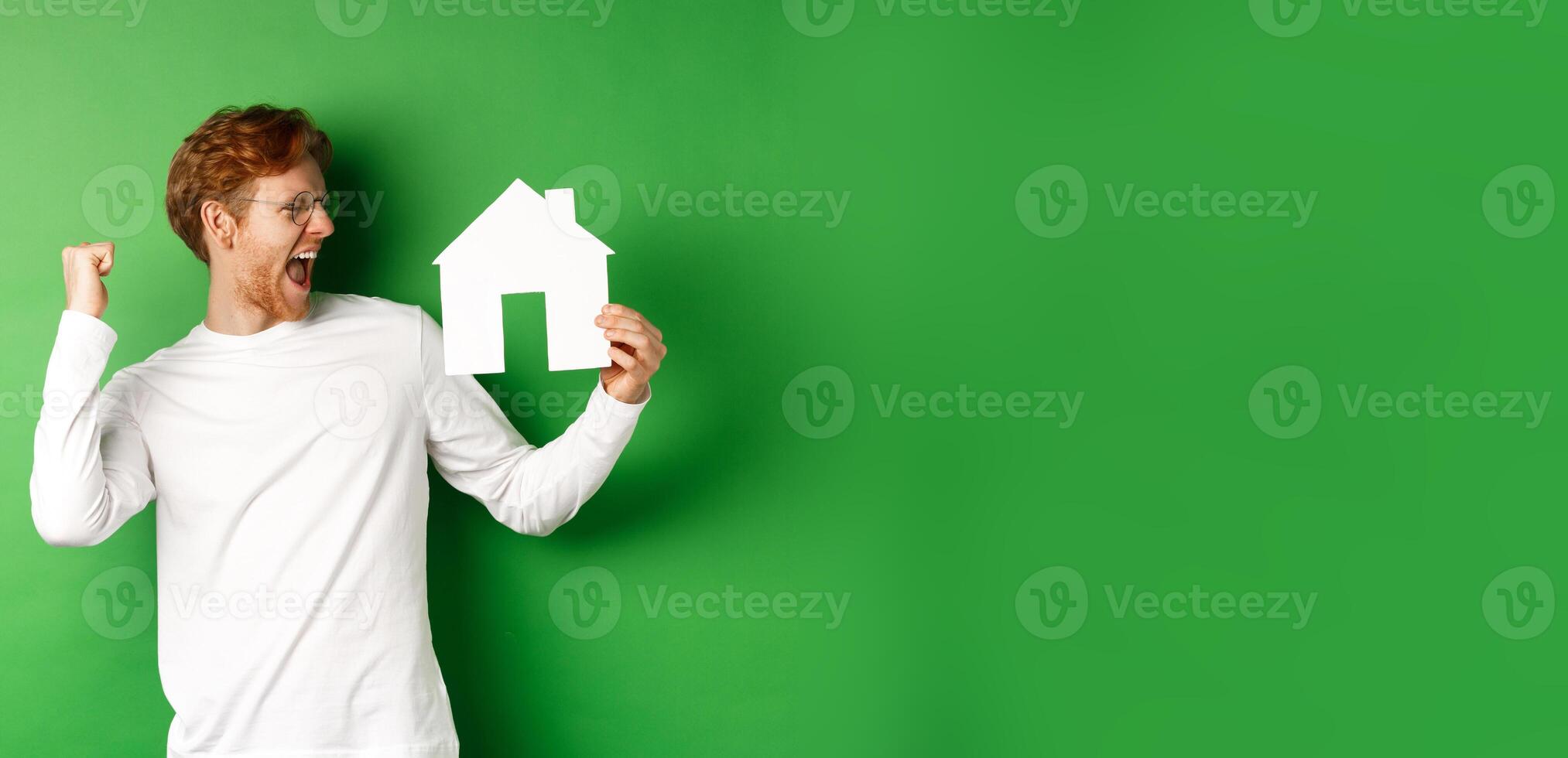 Real estate. Cheerful redhead man in glasses celebrating, buying a house, showing paper home cutout and scream with joy, standing over green background photo
