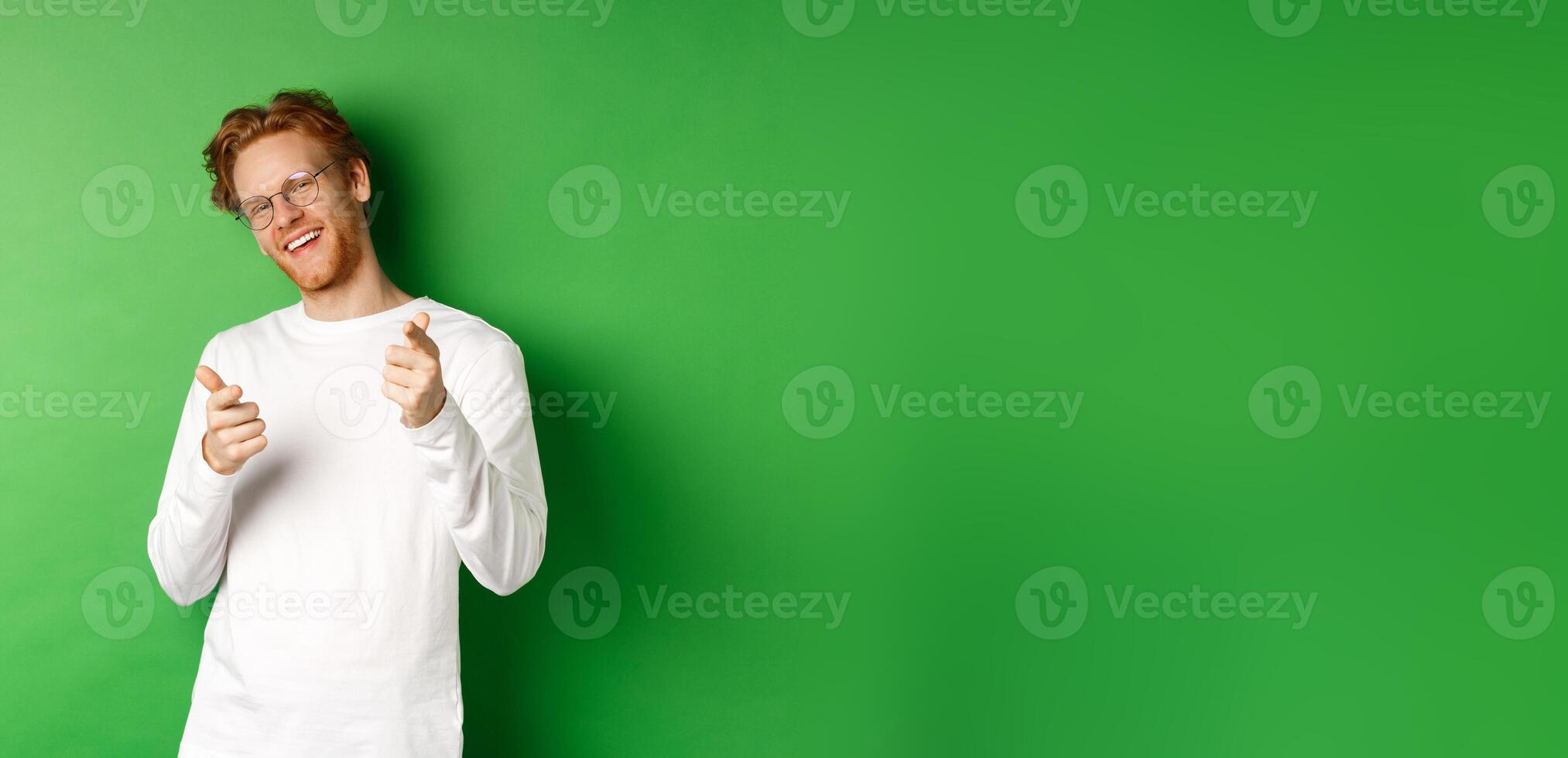 Cheeky young man in glasses praising and congratulating you, pointing fingers at camera and smiling, standing in glasses and white long-sleeve against green background photo
