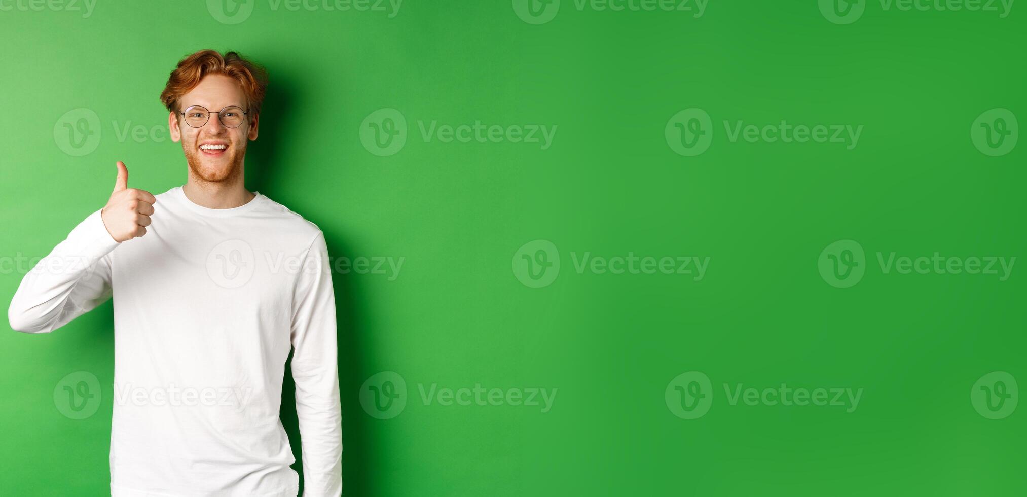 Handsome young man in glasses with red hair, showing thumbs-up and smiling satisfied, praising good eyewear store, standing over green background photo