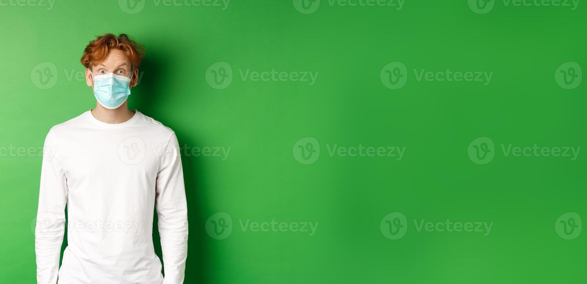 Covid-19, social distancing and lifestyle concept. Young redhead man wearing face mask during coronavirus pandemic, looking surprised at camera, green background photo