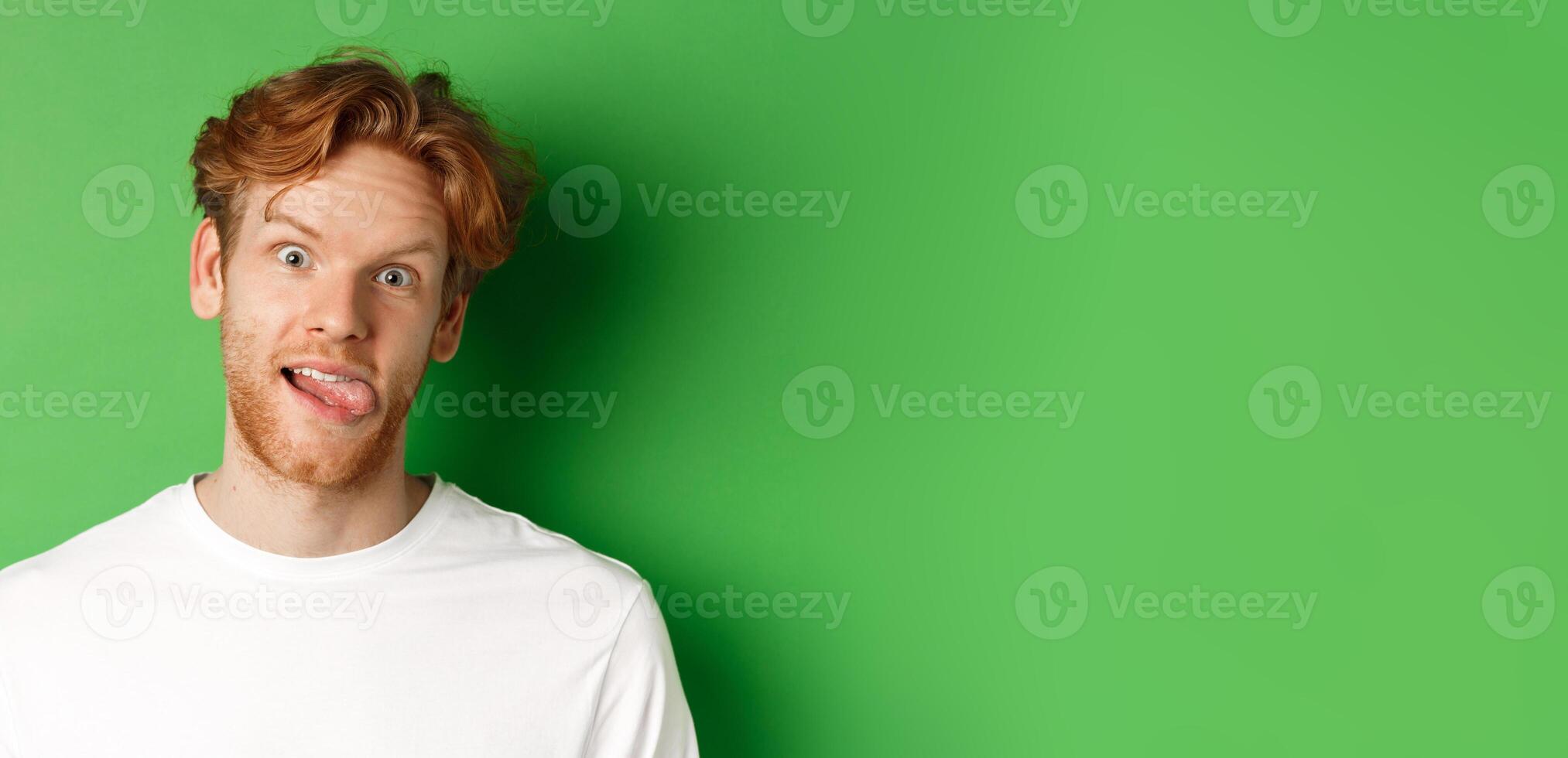 Emotions and fashion concept. Close up of funny redhead man showing silly faces, sticking tongue and staring at camera, standing over green background photo