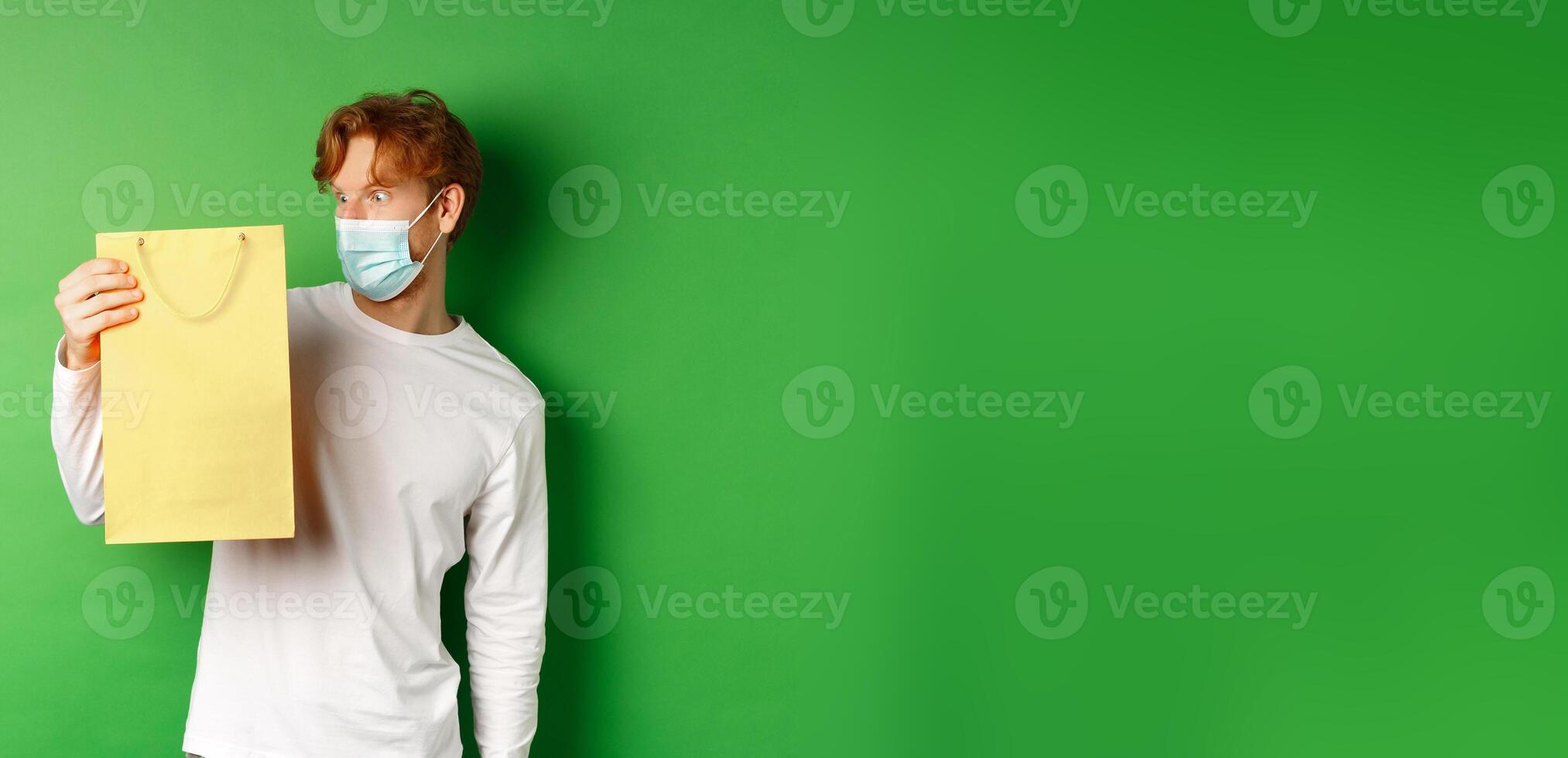 Surprised redhead guy receiving a present, looking at shopping bag with curious face, standing over green background in face mask from covid-19 photo