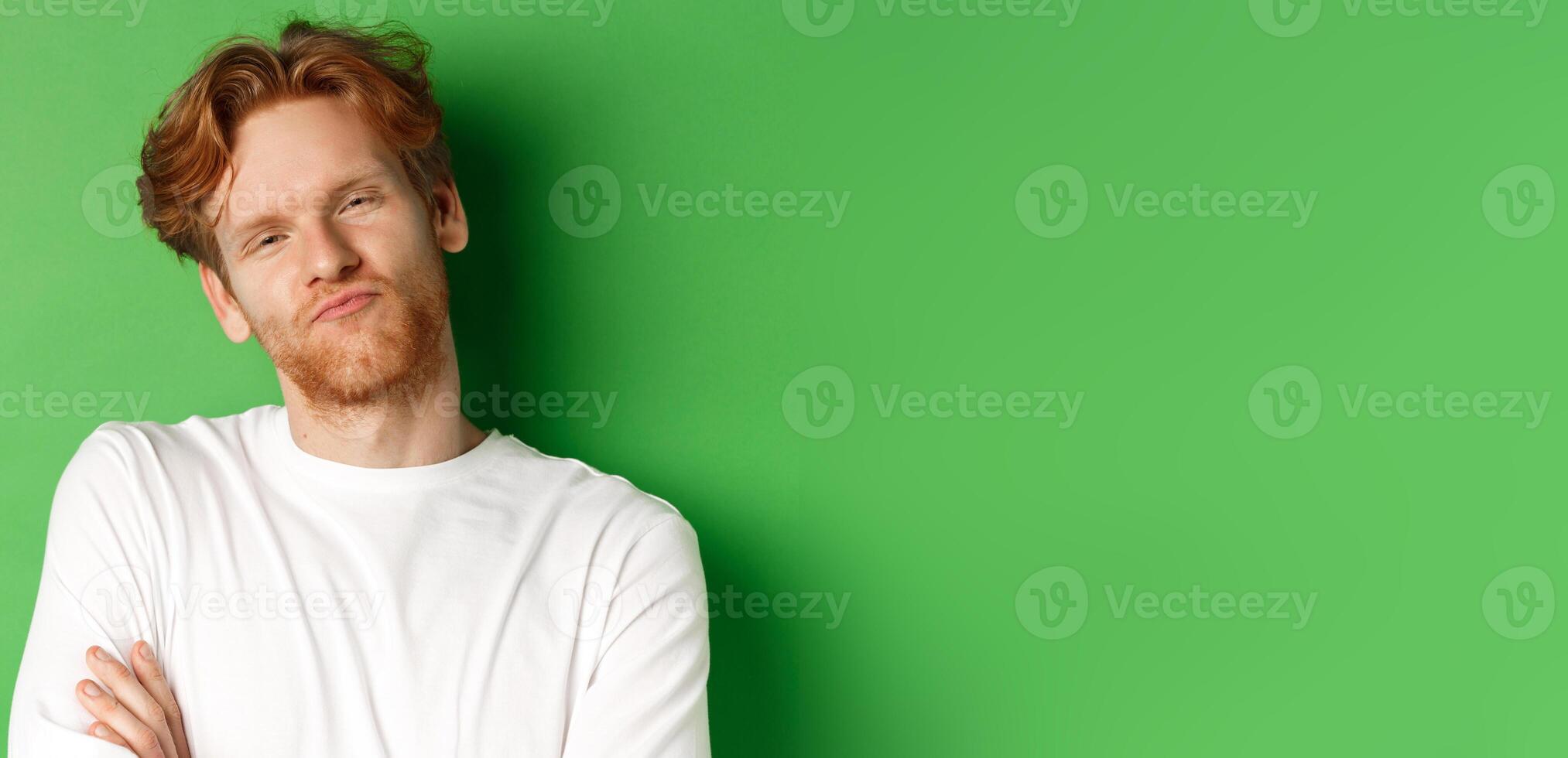 Emotions and fashion concept. Close-up of skeptical young man with red hair and beard pucker lips, looking at camera as waiting for something, standing over green background photo