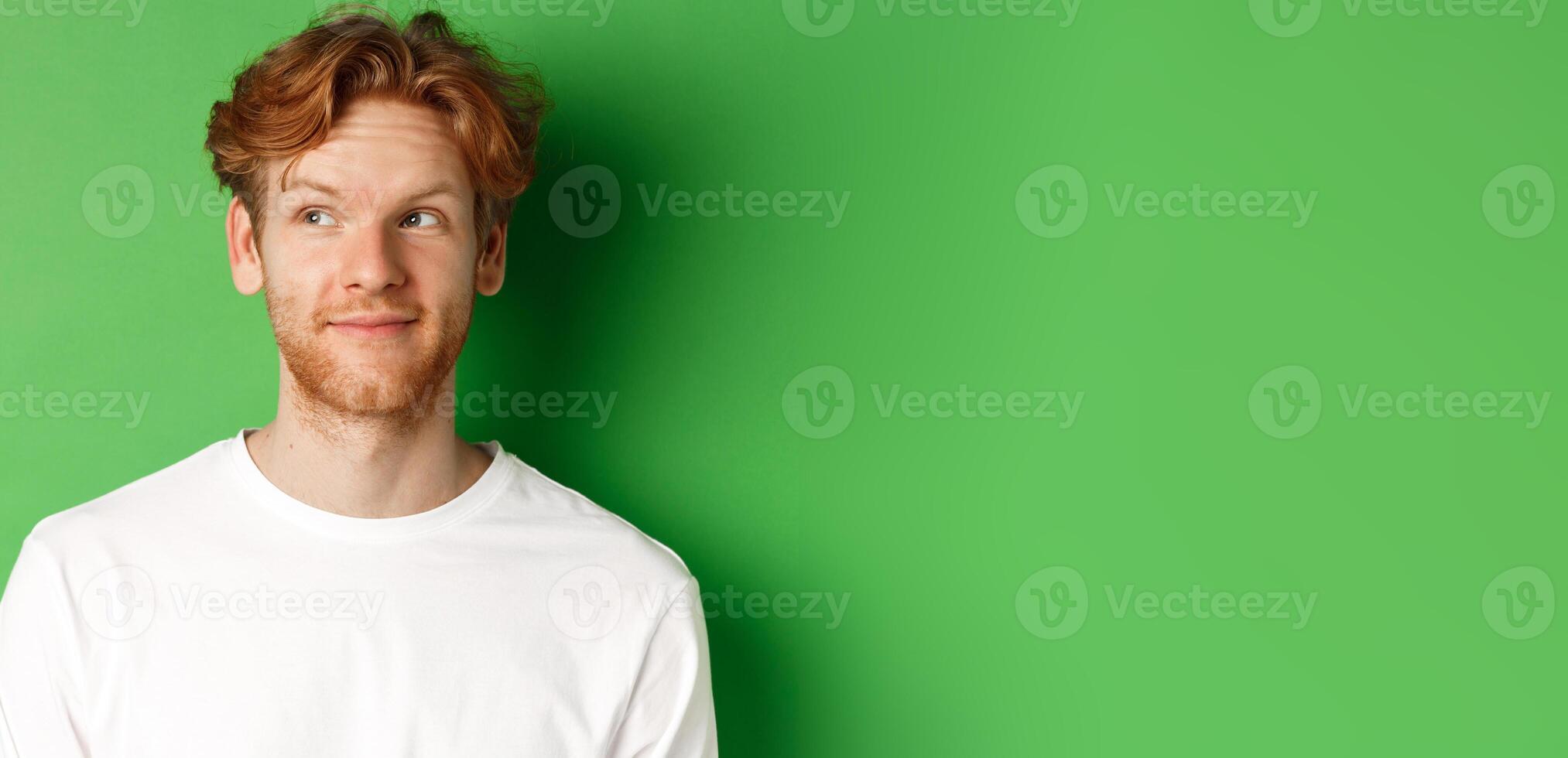Emotions and fashion concept. Close-up of handsome redhead male model with beard and messy haircut, looking left and smiling, green background photo