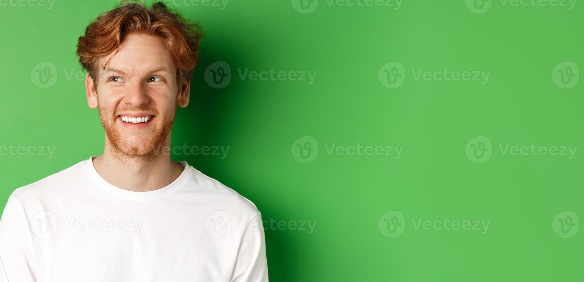 Headshot of happy redhead man with beard wearing white long sleeve, looking left at copy space and smiling, standing over green background photo