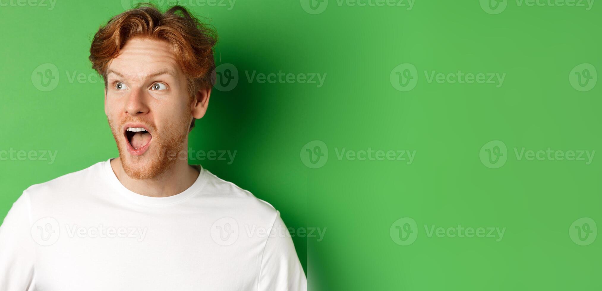 Close up of surprised and impressed redhead man checking out promotion offer, looking left with dropped jaw, standing over green background photo