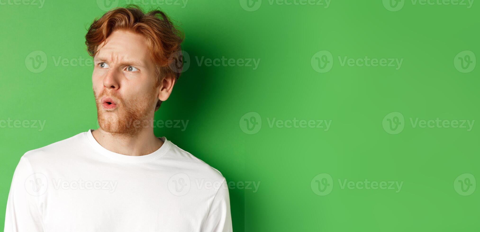 Confused redhead man staring left and looking worried, standing against green background photo