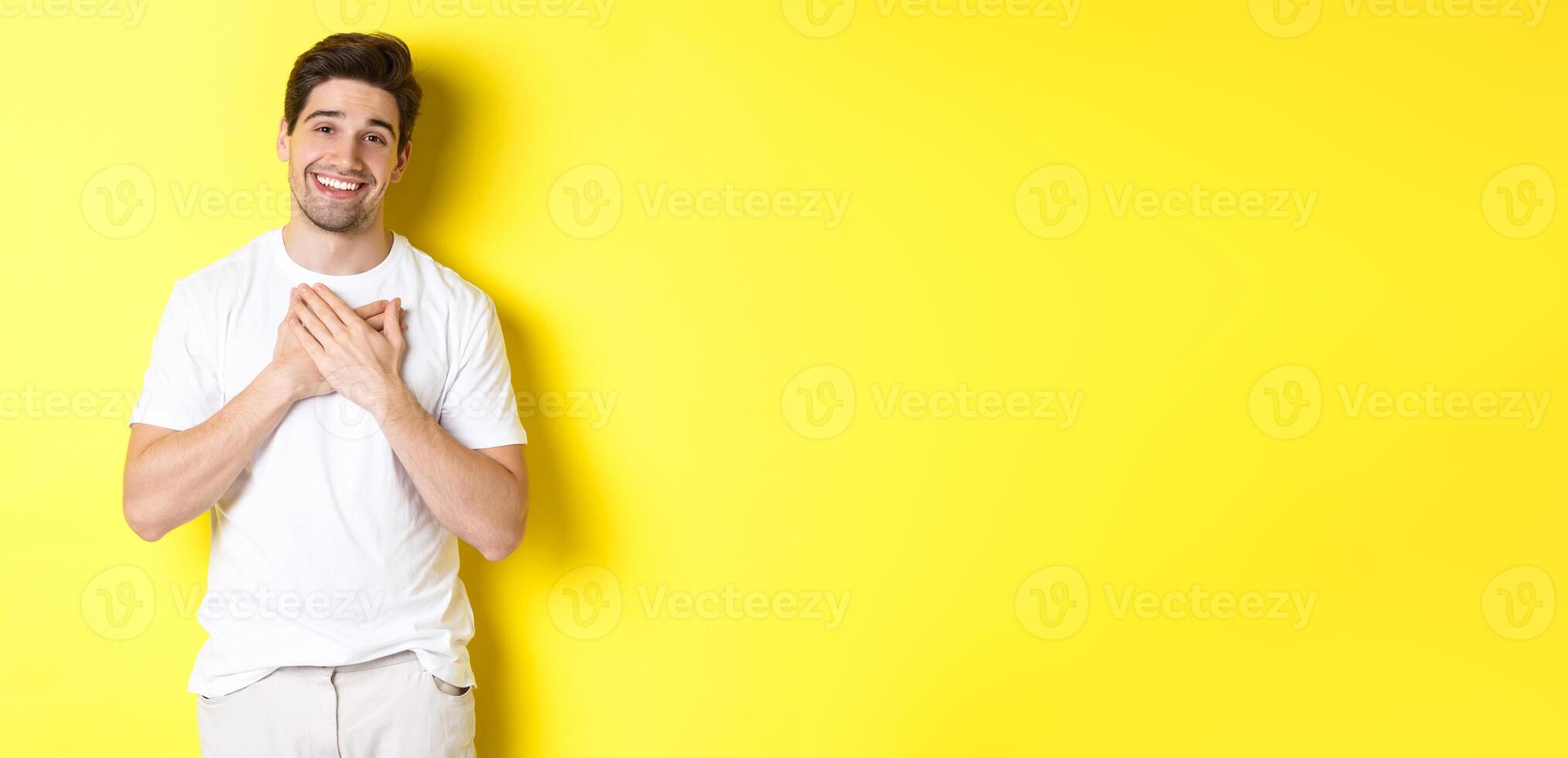 Image of grateful handsome guy in white t-shirt, holding hands on heart and smiling pleased, express gratitude, thanking for something, standing over yellow background photo